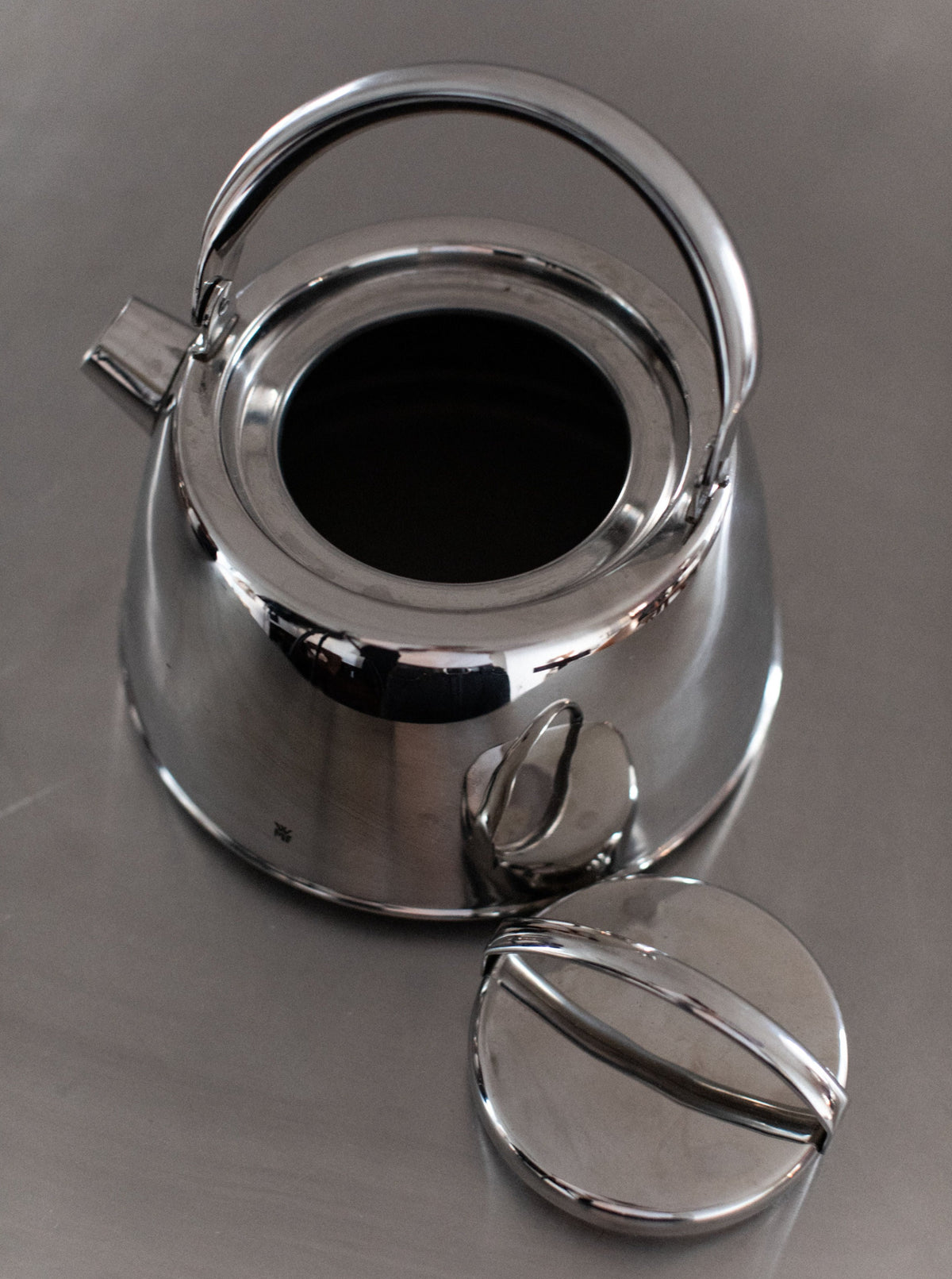 A polished stainless steel kettle with an open lid sits on a reflective surface, showcasing its aesthetic appeal. The Out For Lunch Chrome Teapot's lid is placed to the side, revealing the interior. It features a smooth, shiny finish with a sturdy handle arc over the top, enhancing any dining experience.