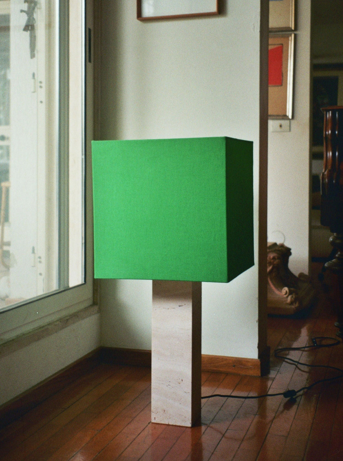 The Roma I Lamp Squared White Travertine, Green Linen Limited Edition by Nassi Lamps sits on a wooden floor next to a large window. Its squared green linen lampshade on a rectangular base contrasts with a white wall and various framed artworks, embodying elements of modernism.
