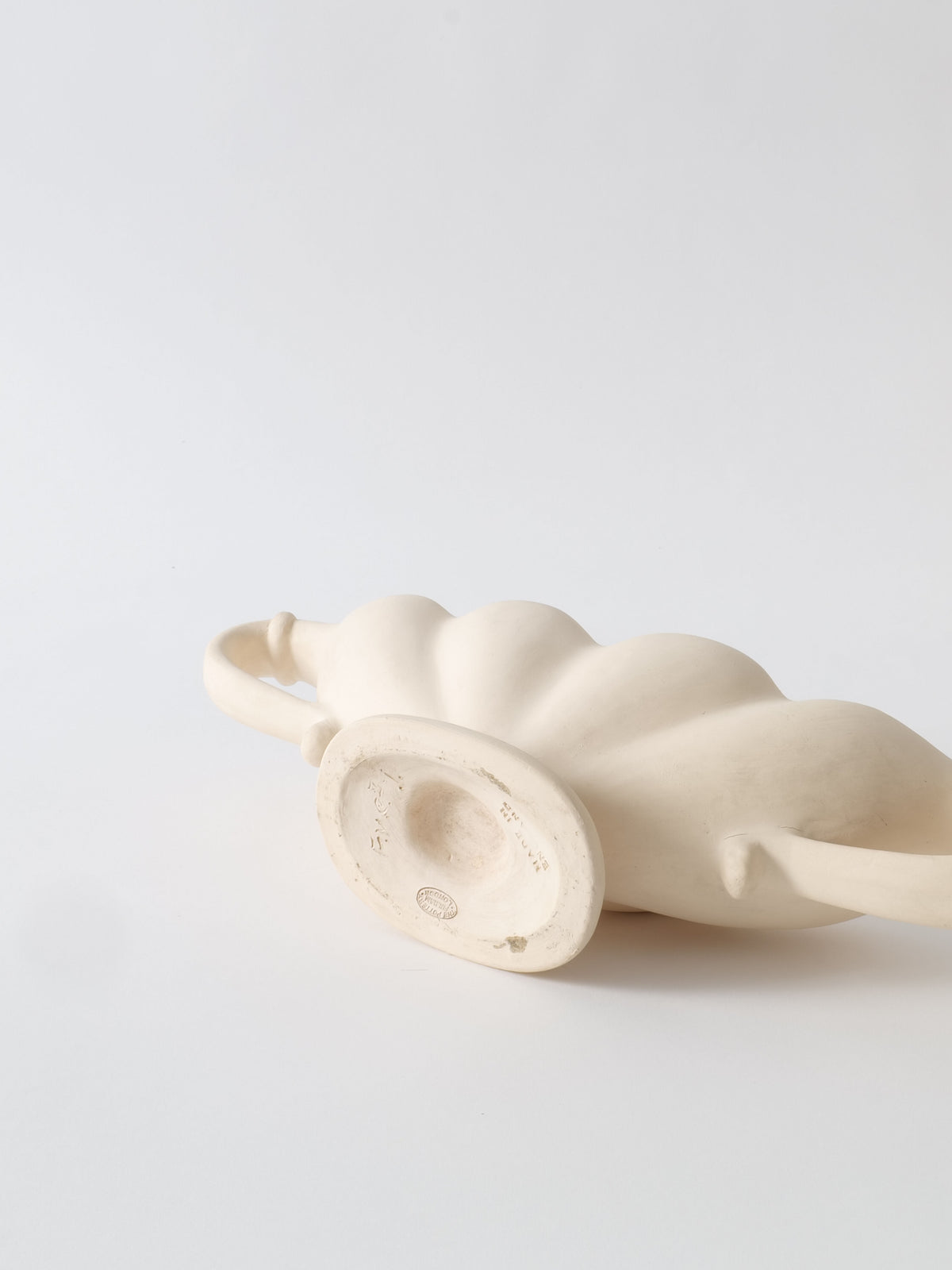 A beige, curvaceous ceramic vase from the 1930s, known as the Pleated Vase and crafted by Collection apart, is made from Devon clay with a small handle, presented against a white background. The Fulham Pottery-attributed vase lies sideways with its round base facing the viewer, showcasing a subtle texture and minimalist design.