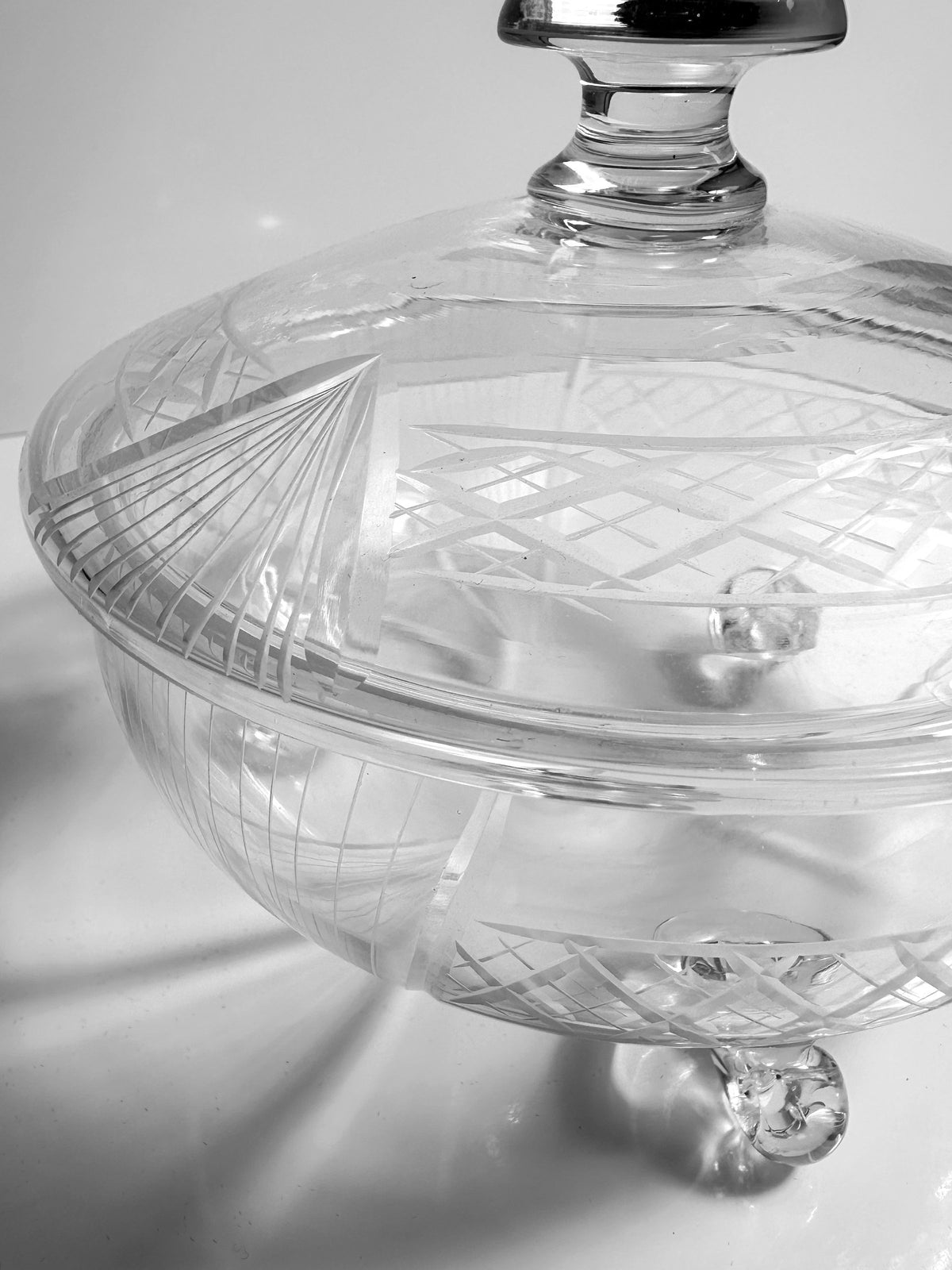 A close-up of the *Art Deco Glass Bonbonnière - 1930* from *Dodo Vintage* showcases its transparent glass body adorned with intricate etched patterns and a matching lid. The design embodies Art Deco elegance with geometric shapes and refined lines, contributing to its sophisticated appearance. This handmade piece is beautifully displayed on a white surface.