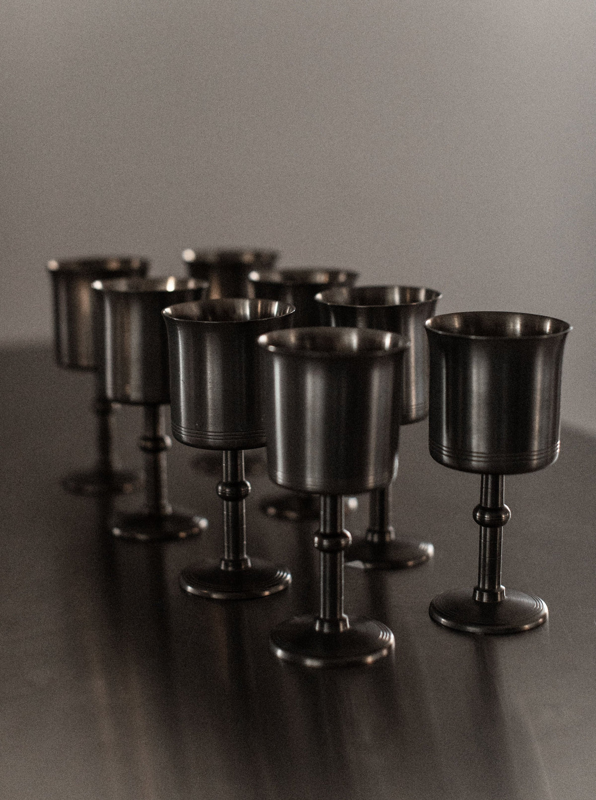 A set of eight Art Deco Cups by Out For Lunch, each with a slightly tarnished appearance, is arranged in a staggered formation on a dark, reflective surface. Their design radiates timeless elegance, while the blurred background enhances the image's moody, vintage feel.
