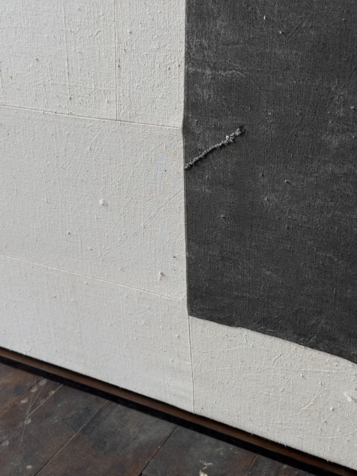 A close-up of a textured surface featuring a large off-white area next to a smaller dark gray area, reminiscent of Chinese cotton patchwork. Both surfaces have a rough, slightly uneven texture, with the scene partially showing a wooden floor at the bottom of the image. This intricate design is part of the (a-tent), 2024 Framed Hand Loomed Tapestry by Bec Kirby.