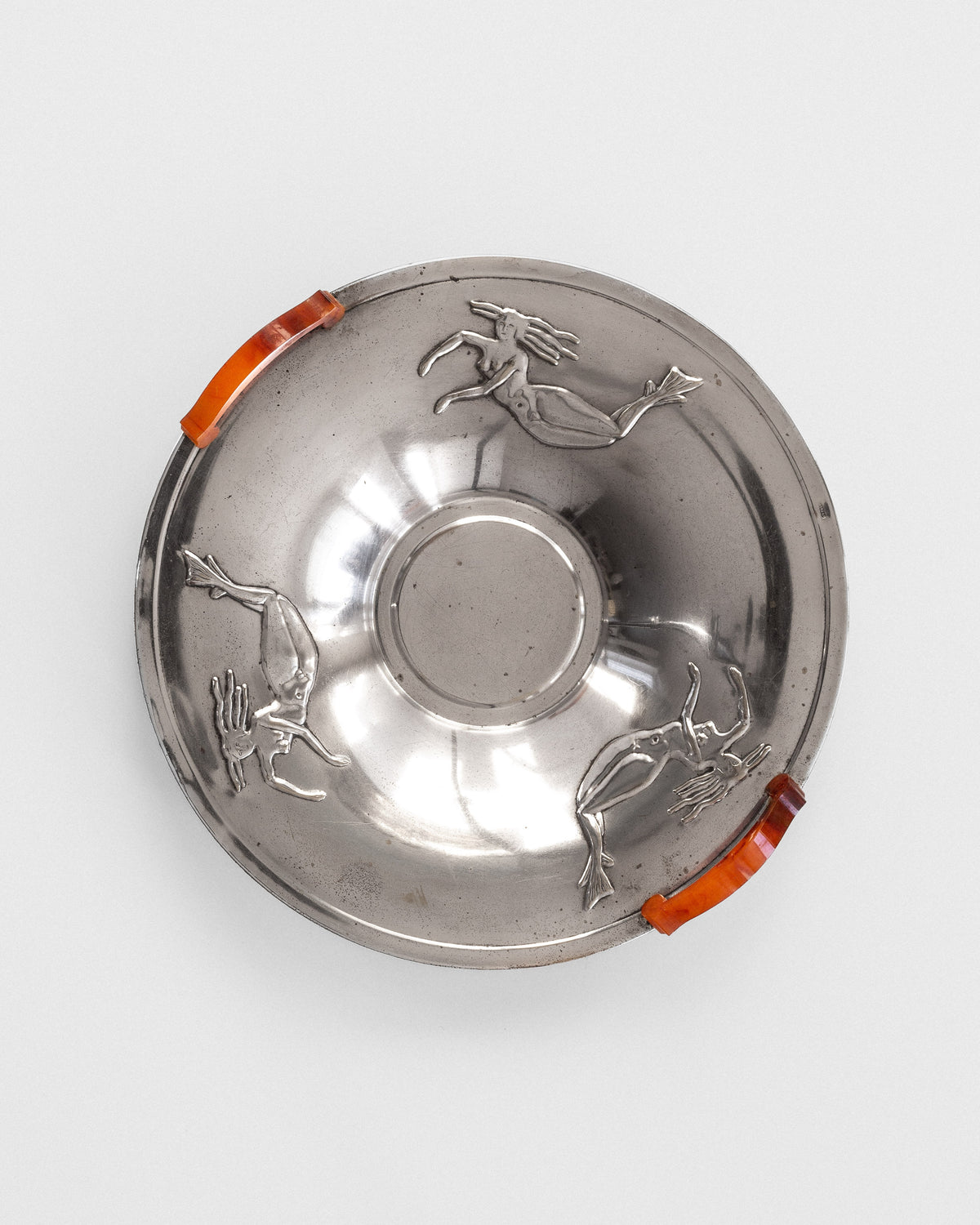 This Decorative Bowl with Mermaids from the 1930s by Spigel features a round, silver-plated design with two orange glass handles. Embossed figures of mermaids adorn its exterior, embodying the elegance of the Swedish Grace movement against a simple white background typical of the Art Deco period.
