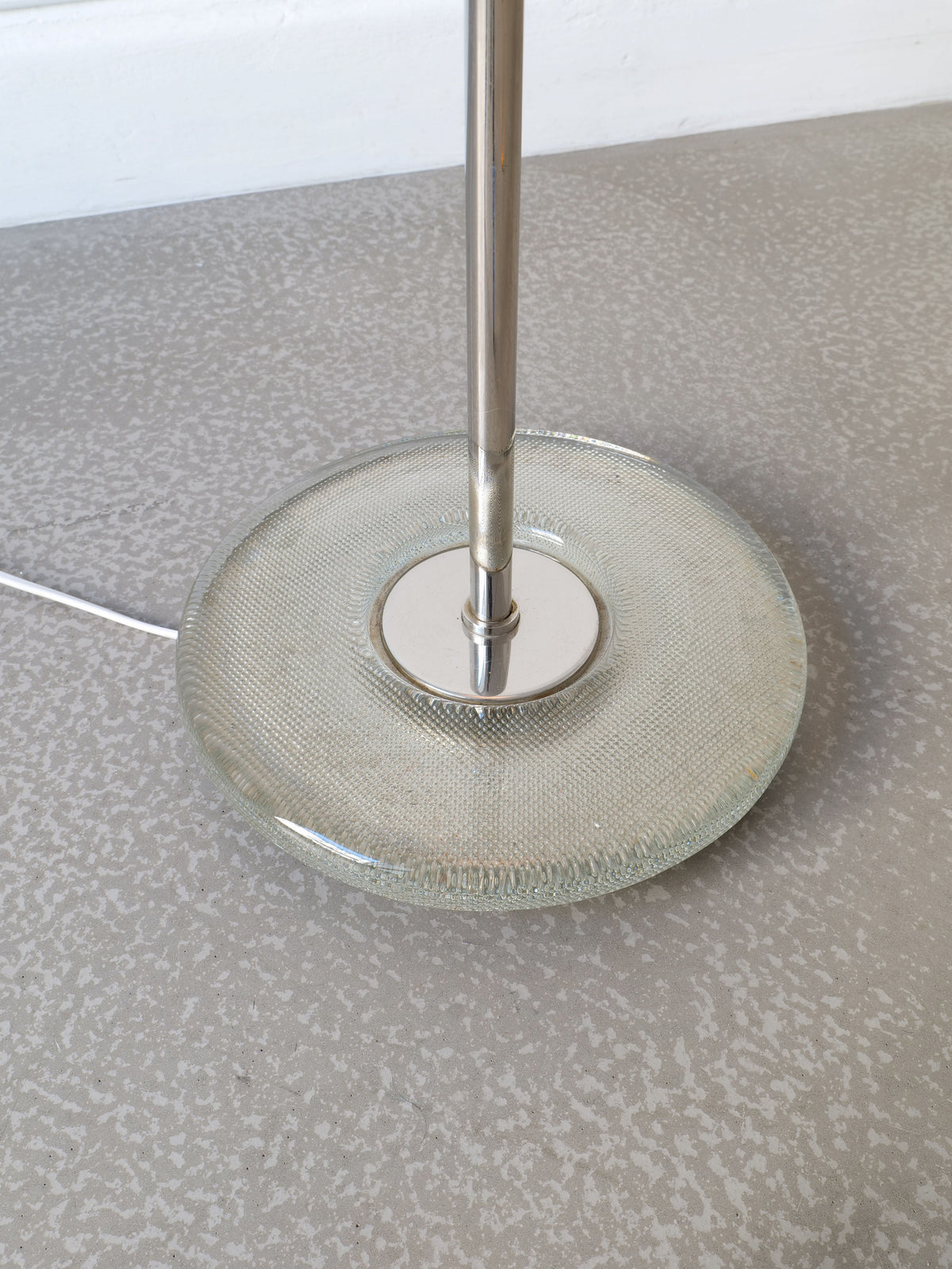A close-up of the Deco Floor Lamp Harald Notini 1940s by Collection apart shows its round, textured glass base with a central metal pole, standing on a speckled grey floor and a white power cord extending from the base.