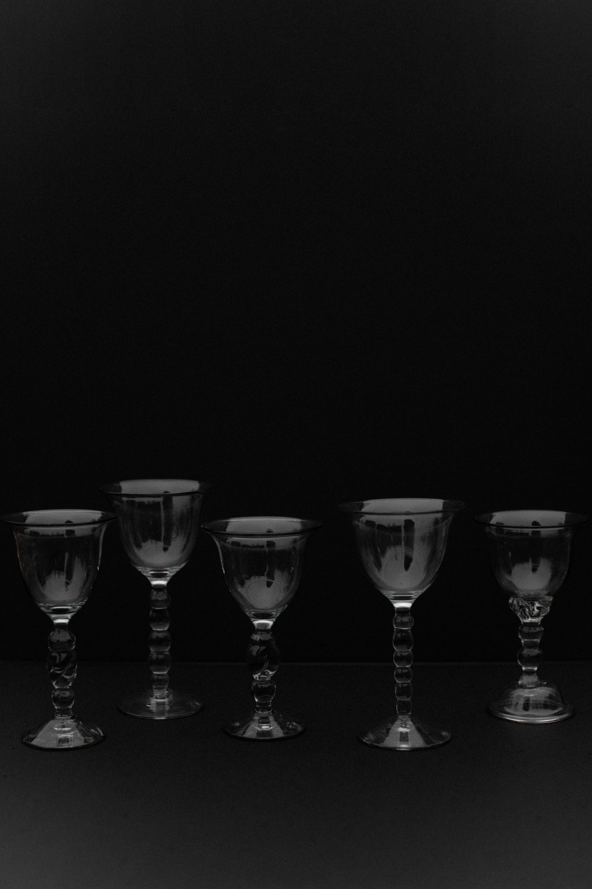 Five handmade art glasses from Out For Lunch are symmetrically arranged against a black background, casting subtle reflections. Their exquisite craftsmanship and varying shapes and sizes highlight their unique beauty as decorative accents.