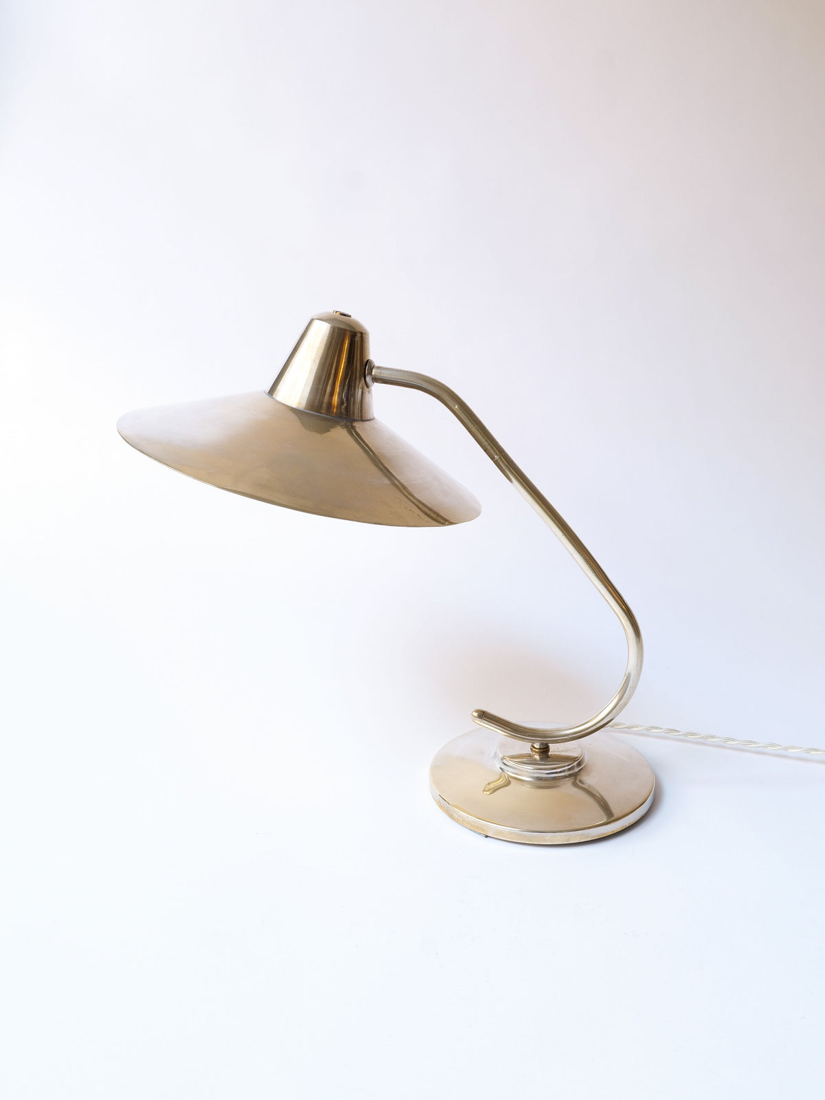 A sleek, modern Collection apart Silver Brass Desk Lamp features a silver-plated brass design with a conical shade and a curved stem. The lamp has a round base and a white cord extending from the back, set against a plain white background, channeling midcentury elegance.