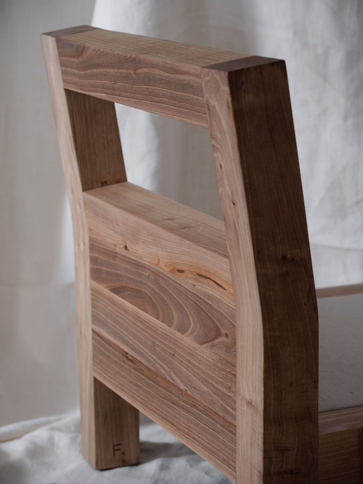 A close-up of the "Forma" Armchair by FEBRERO SHOP shows a solid light brown wooden backrest and seat with a smooth, natural grain pattern. Against a soft linen fabric backdrop, it highlights its simple, rustic design.