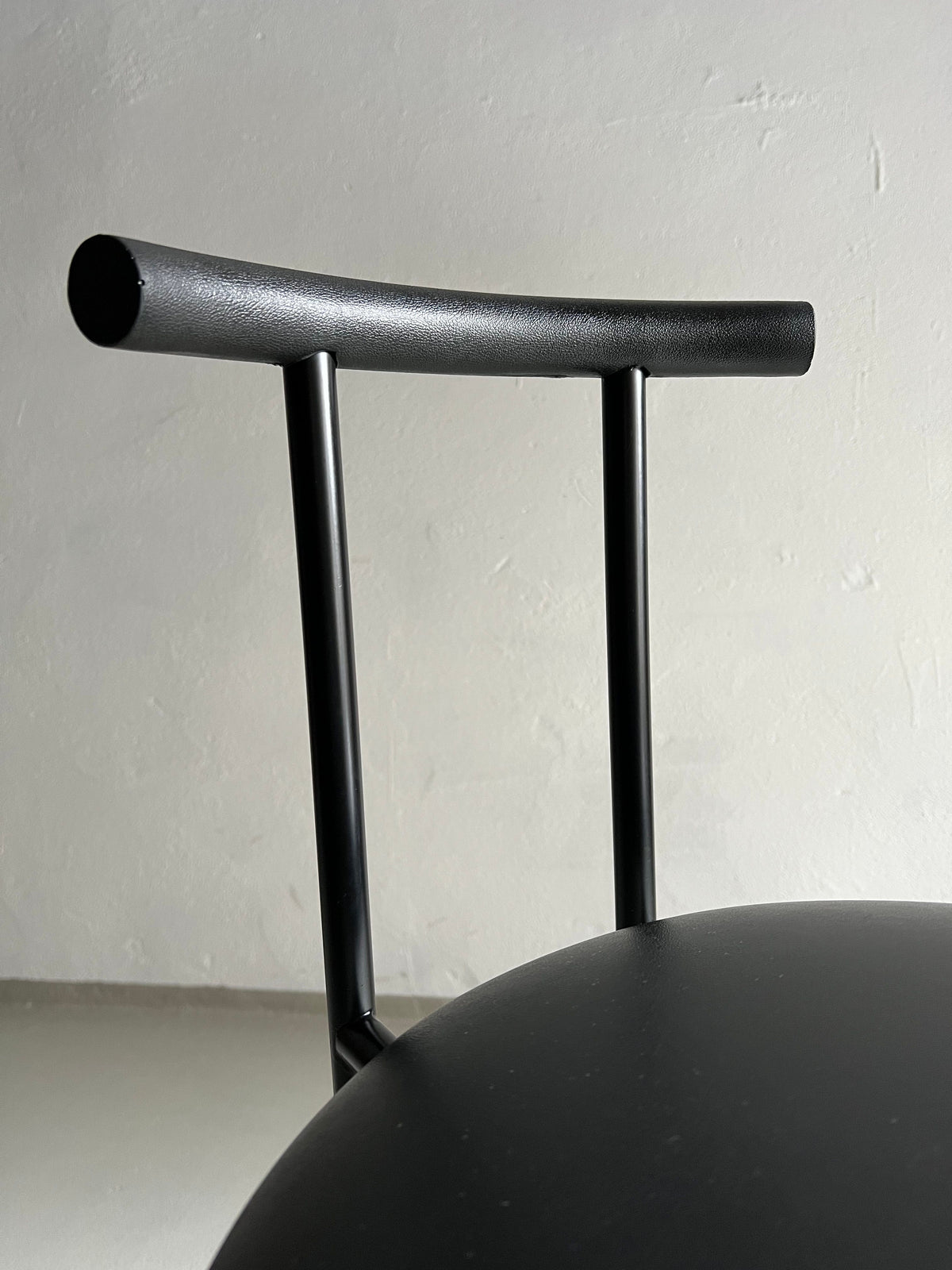 Minimalist black chair with a cylindrical backrest and faux leather upholstery. The chair is set against a light grey wall, highlighting its sleek and simple design. The slightly curved backrest complements the round, smooth seat. This is the 4 Postmodern "Tokyo" Chairs by Rodney Kinsman | Italy | 1980s from Veter Vintage.