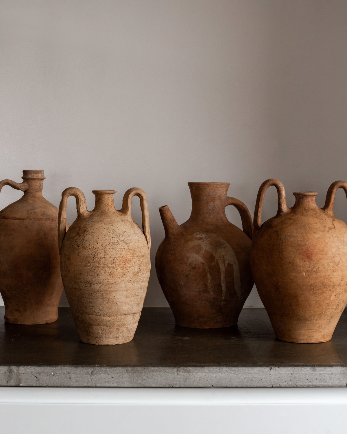 Set against a white backdrop on a gray surface, four rustic Spigel Early 20th Century European Ceramic Pitchers flaunt their elegant handles. Varying in shape and size, these hand-crafted brown pieces feature a weathered, textured finish that highlights their unique decorative charm.
