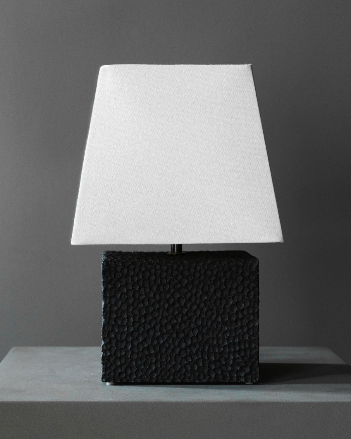 The Akune Ceramic Lamp - Number Five by Studio OSKLO showcases post-Bauhaus brutalism with its textured black rectangular base and simple white square shade, displayed on a gray surface against a dark gray background.