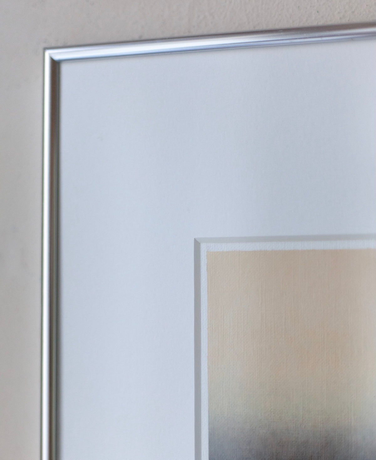 A close-up of a corner of Rebecca von Matérn's "Untitled, 2024" framed acrylic painting on a light-colored wall. The frame is made of aluminum with a narrow border, showcasing soft, blended hues of beige, gray, and off-white in the visible part of the artwork.