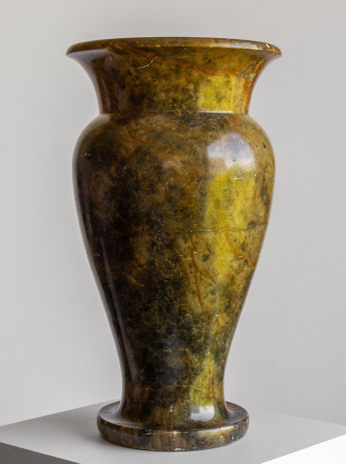 A large Neoclassical Stone Vase by Spigel, boasting a flared rim and an urn-shaped design from the early 1900s, with a rich mix of dark green, brown, and yellow hues on its tall polished marble surface, sits on a white pedestal against a light grey background.