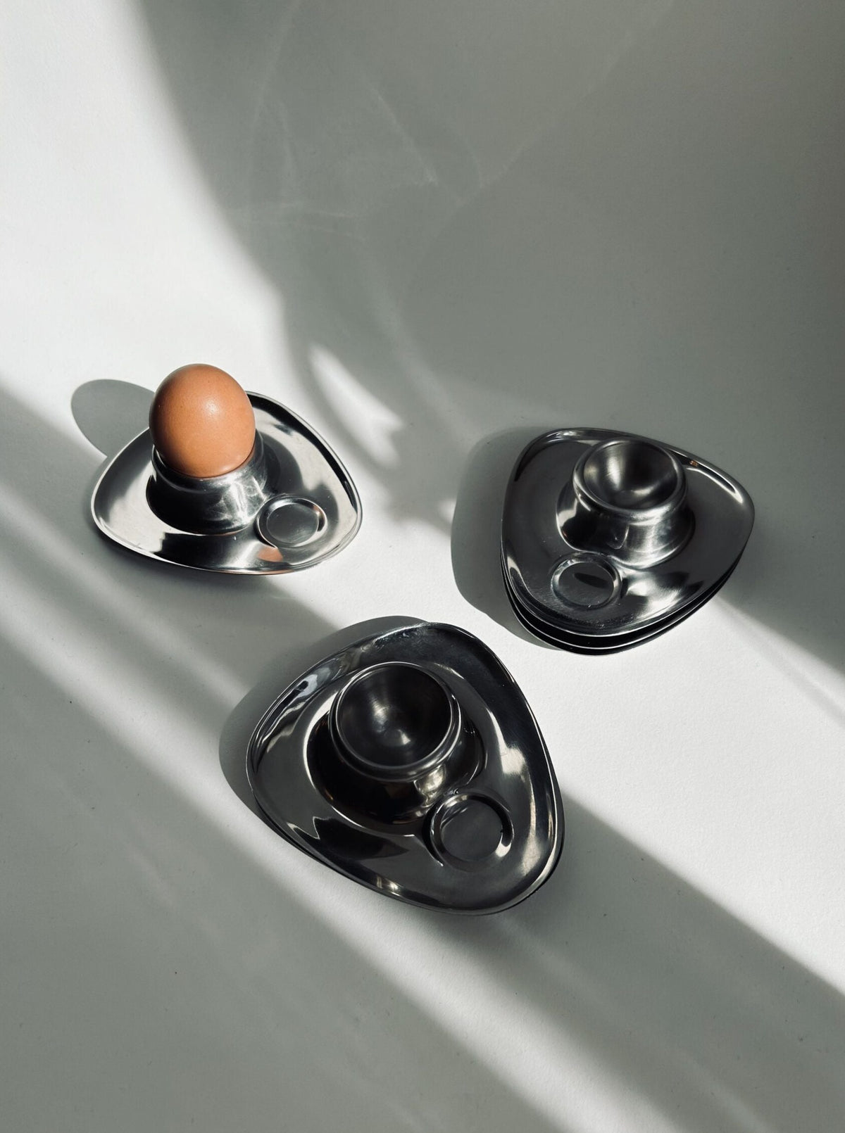 A trio of sleek, triangular egg holders from the Bauhaus Stainless Steel Egg Cups Set of 6 by Flock Studio are arranged on a white surface. One holder cradles a brown egg, while the others remain empty. Soft shadows stretch across the scene, adding depth and creating an elegant display reminiscent of vintage mid-century design.