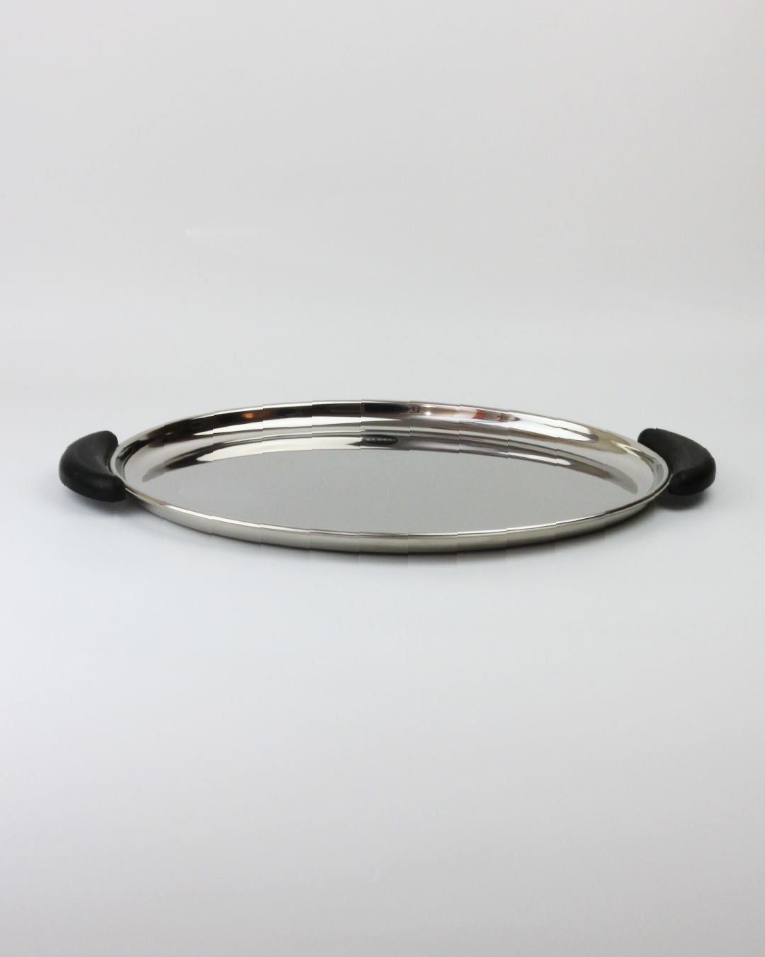 A round stainless steel serving tray with two black handles on either side, placed on a plain white surface. The tray, reminiscent of mid-20th-century craftsmanship, features a smooth, reflective surface and a slightly raised edge. Perfect for showcasing the elegant "Bombè" Alessi Tea/Coffee Set from Boga Avante Shop.