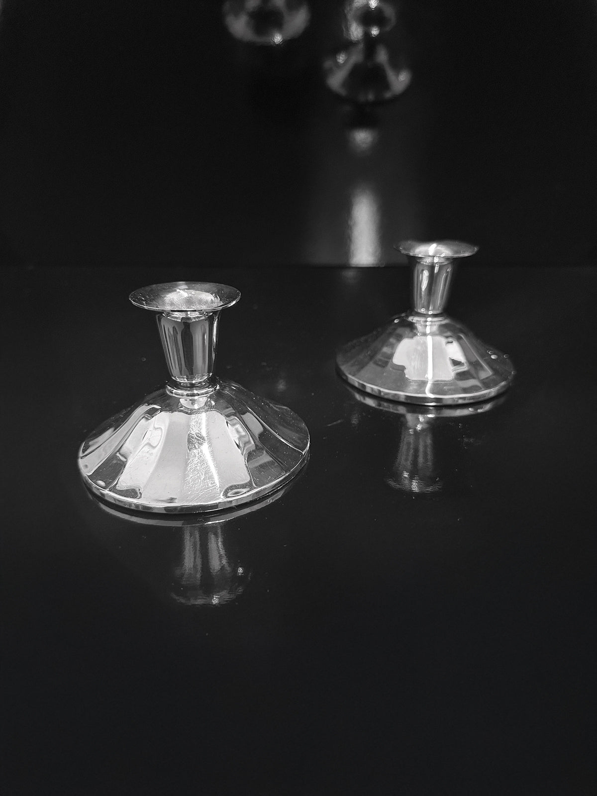 A pair of Silver-Plated Art Deco Candleholders from 1930, by Dodo Vintage, with reflective, fluted bases are positioned on a dark, glossy surface, capturing their reflections. The dark backdrop accentuates the gleaming and polished appearance of these exquisite candlesticks.