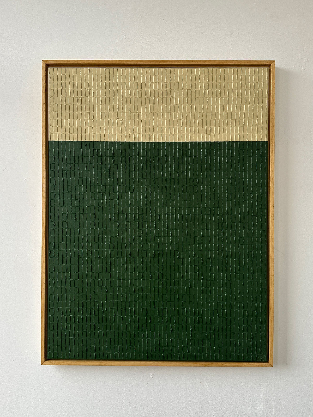 The "Buff Verte" oil on canvas by Roi Elmaliah showcases a minimalist design with two horizontal sections: a wide dark green lower portion and a narrower beige upper portion. The textured canvas is elegantly framed in light wood against a plain white wall, creating the serene ambiance reminiscent of an art gallery in Amsterdam.