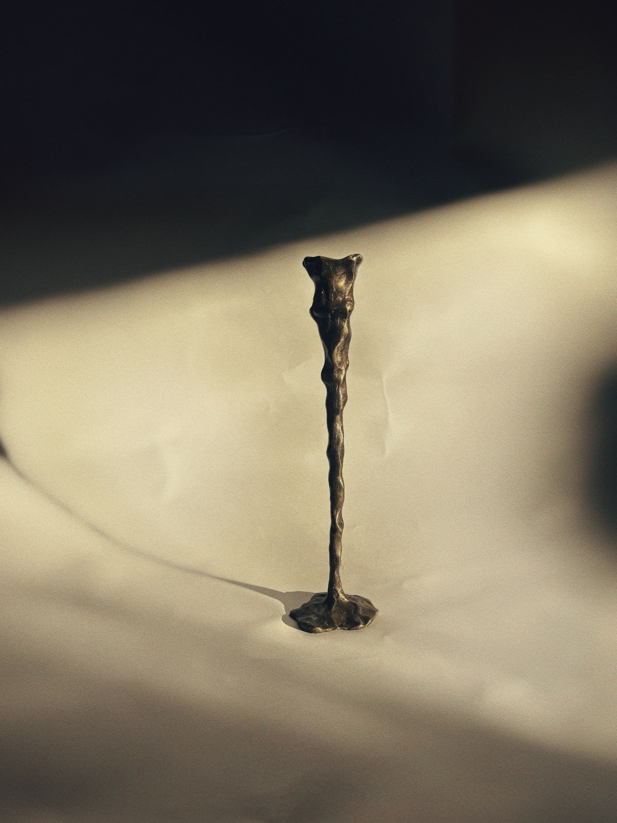 A slim, abstract creation by Huerto mimics a Bronze Candle Holder and stands on a textured surface, casting a shadow. This artist-signed piece is tall and elongated, with a rough, uneven finish, set against a softly lit background that evokes an artistic and contemplative ambiance.