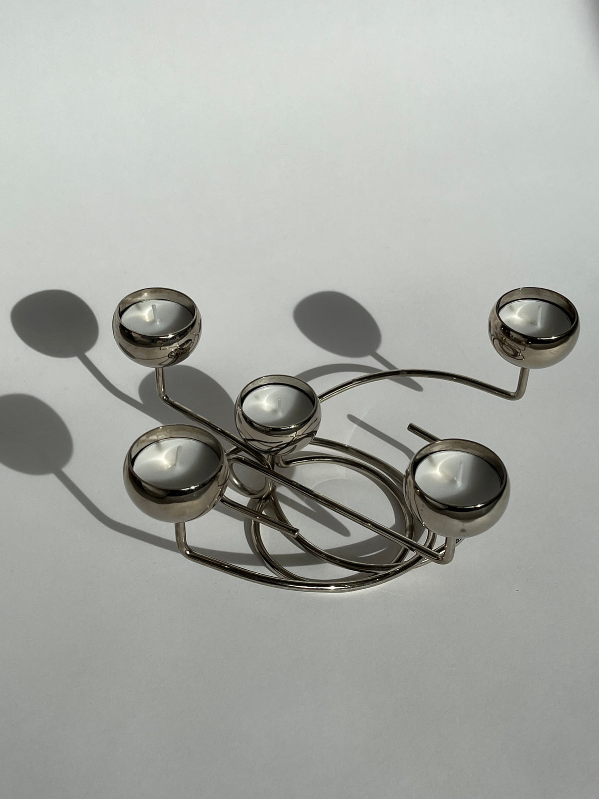The Chrome Candleholder by septembre studios features five round holders in a vintage design, casting soft shadows on a light gray surface. Its intricate metalwork creates an elegant pattern and offers unique angles that enhance its charm.