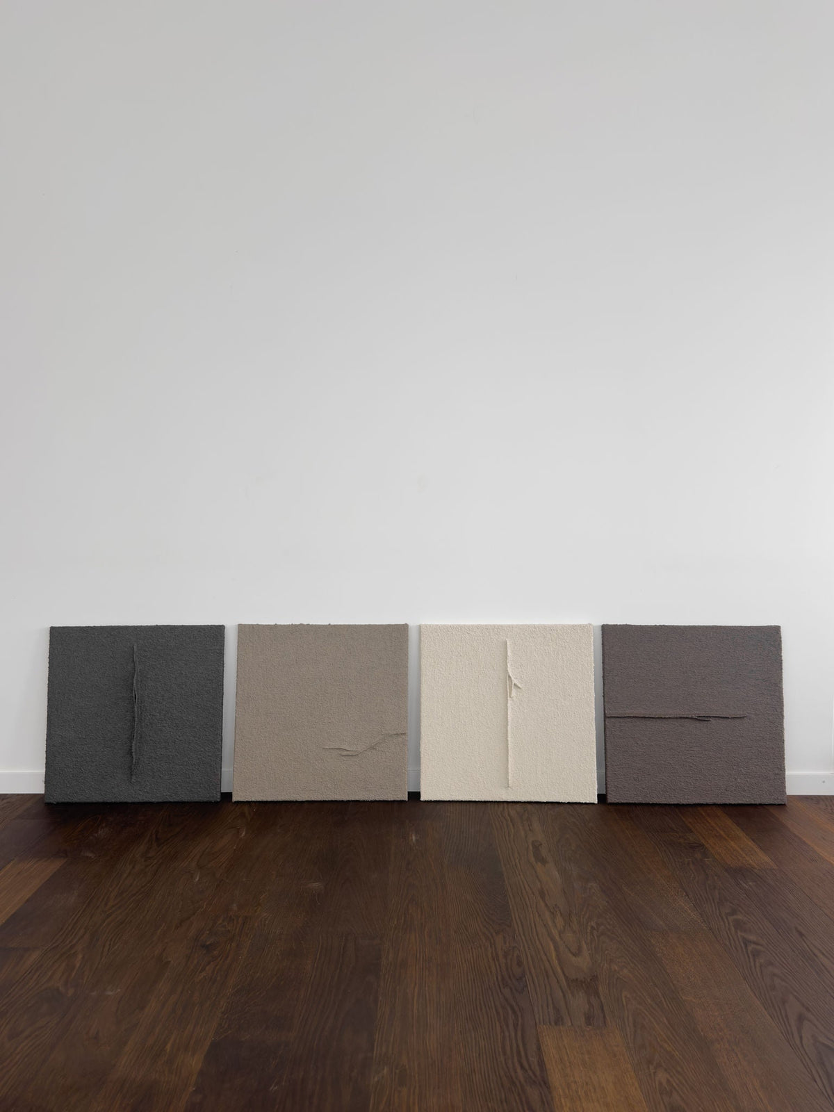 Shadow World 03 by Delal Salazar features four textured abstract canvases in dark gray, taupe, off-white, and brown, created with acrylic and sand. Each unique piece is displayed against a white wall on a dark wooden floor.