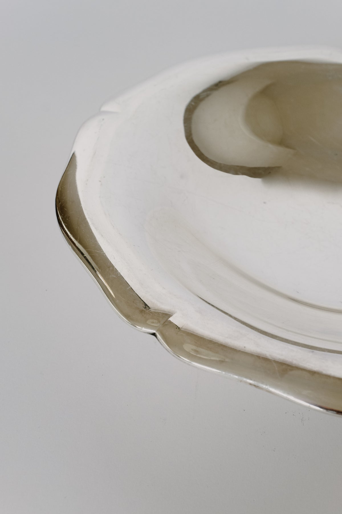 Close-up of a Silverplated Bowl by septembre studios, showcasing its intricate scalloped edge with hints of subtle brownish stains or reflections near the rim. The focus is on the texture and shape against a light gray background, highlighting its vintage appeal and distinctive character.