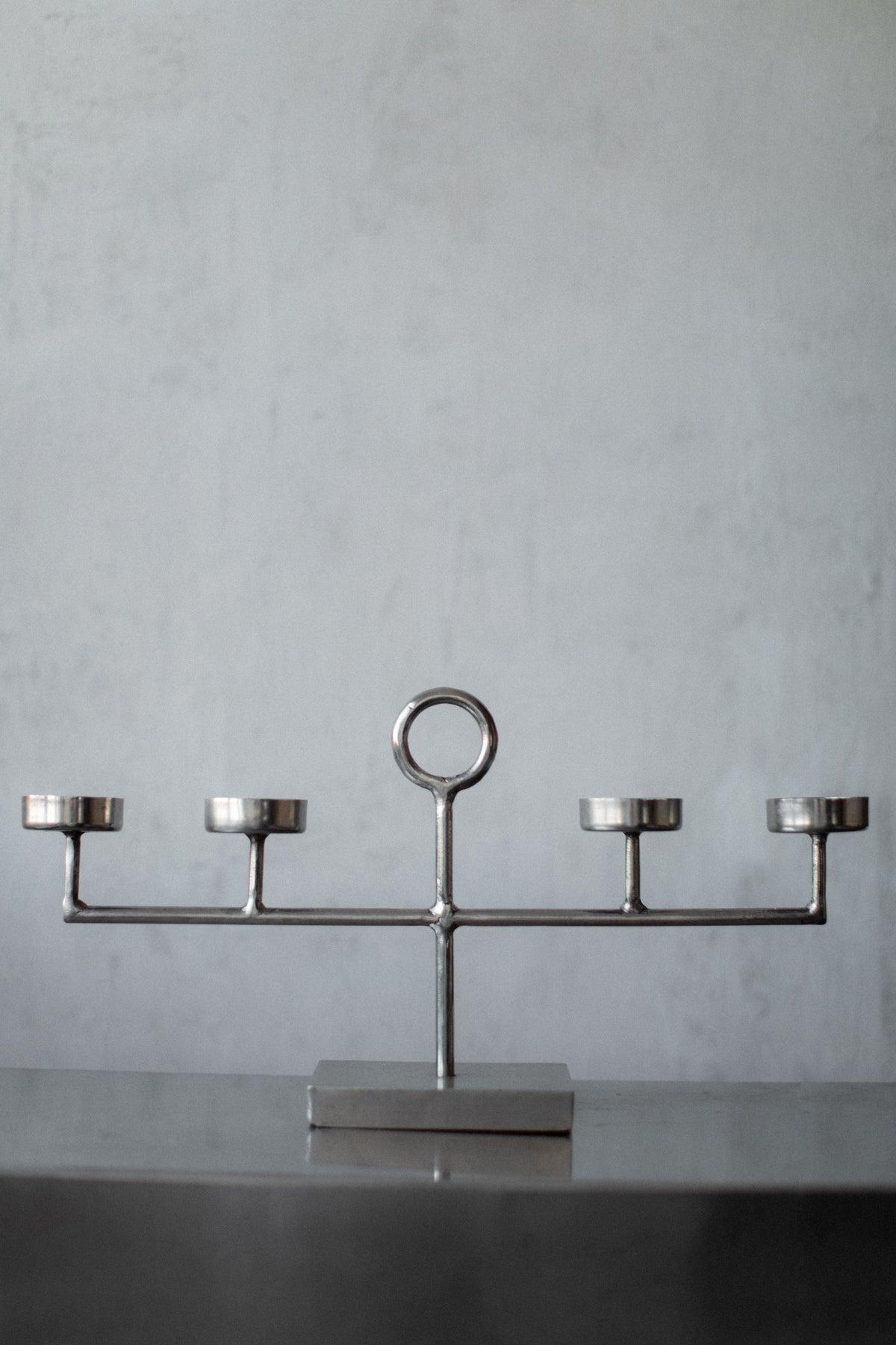 The Out For Lunch Steel Candleholder showcases a minimalist, geometric design with five symmetrically arranged holders and a central circular handle, elegantly resting on a smooth, dark surface against a light gray background.