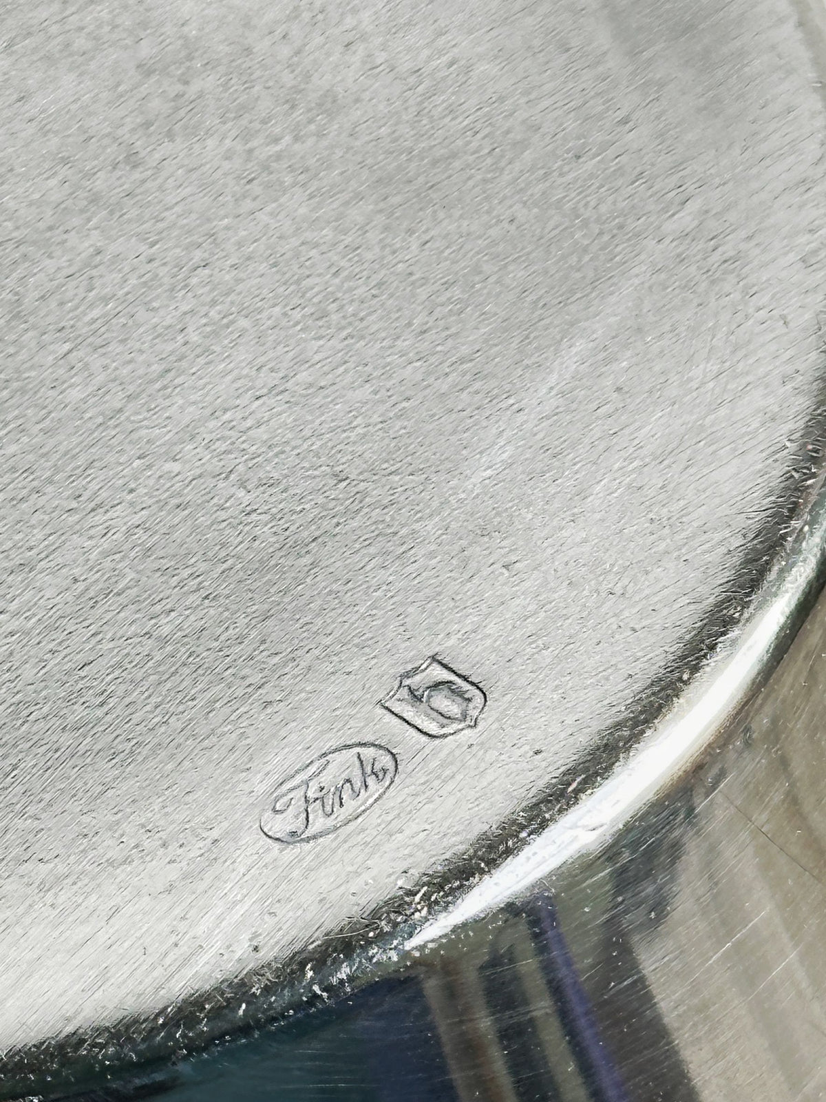 A close-up of a metallic surface reveals two engraved logos: an oval with "Fink" and a shield, representing the Dodo Vintage Heavy Silver-Plated Vase from Fink Germany. The brushed texture and heavy silver plating give it an elegant 1980s jewelry charm at the bottom edge.