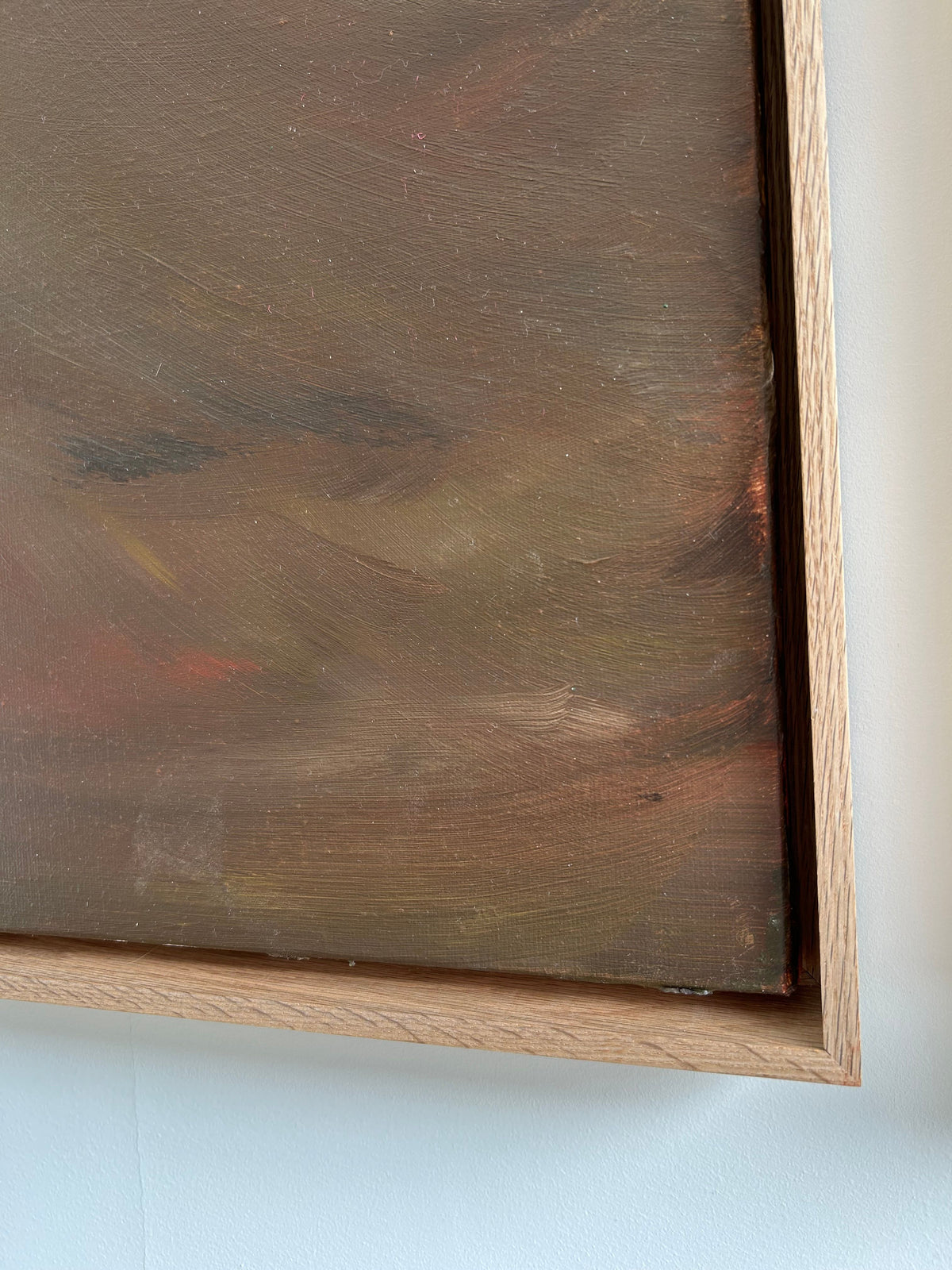 Close-up view of a framed painting from the Terra Incognita (Diptych) - Mixed media on canvas by Mattias Storm, showcasing an abstract, muted palette with shades of brown, green, and hints of red. The wooden frame has a natural finish. The image focuses on a corner of the diptych painting and frame against a white wall.