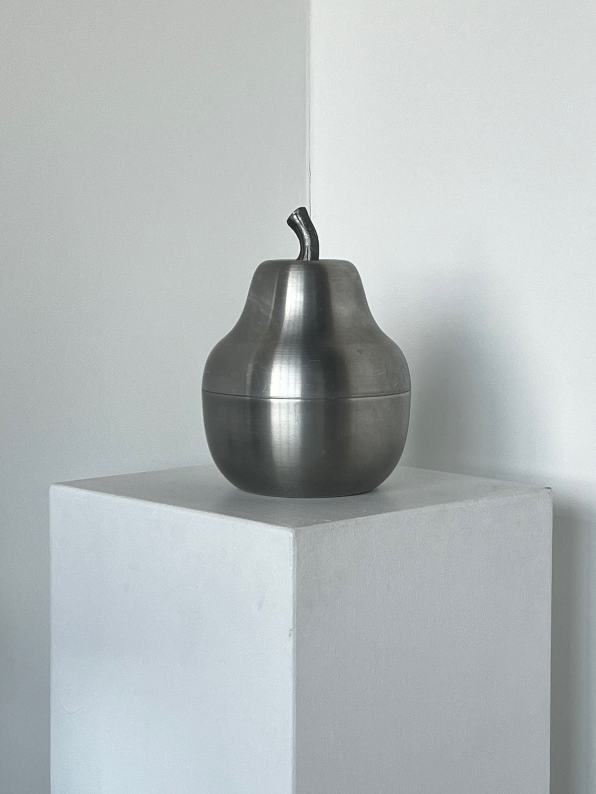 A silver, pear-shaped sculpture resembling the Pear Wine Cooler in Brushed Aluminium by Porter Studio is displayed on a white pedestal against a minimalist backdrop. With its brushed aluminium finish and stylized stem, it echoes the sleek designs of the 1970s condition.