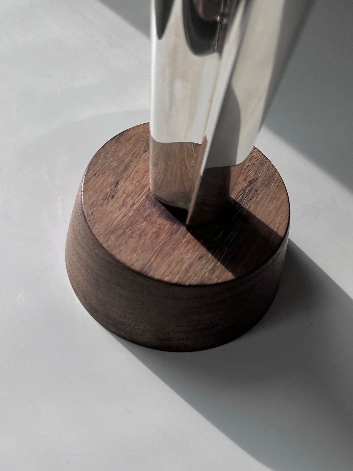 Close-up of a modern sculpture's base, featuring a polished metal structure reminiscent of a Tapio Wirkkala Silver Vase by Médecine, fixed onto a circular wooden platform. The metal's reflective surface contrasts with the wood's natural texture, casting soft shadows on the smooth surface below.