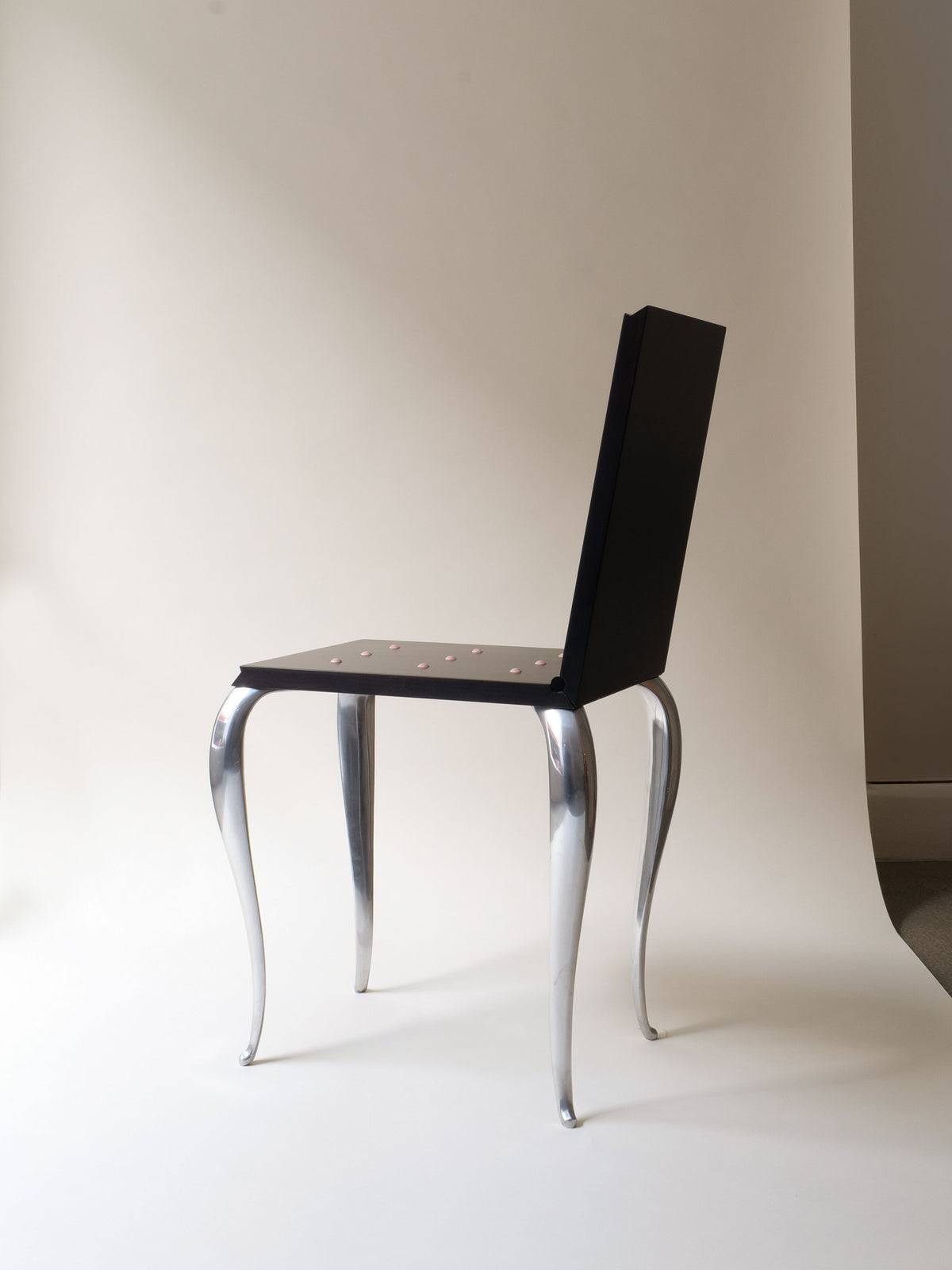The "Lola Mundo Chair" by Philippe Starck (1984) from Collection apart features a black rectangular backrest and seat with four sleek, curved metallic legs, elegantly inspired by Louis XV design, against a neutral studio background.