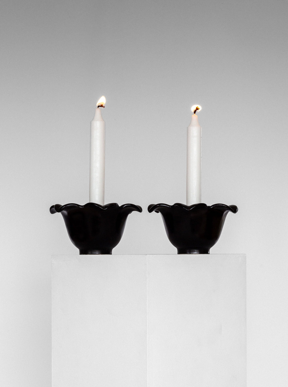 Two white candles are lit and placed in matching black, scallop-edged Spigel Mid-Century Ceramic Candlesticks from Sweden, 1950s. The candlesticks, reminiscent of Swedish Mid-Century Modern studio pottery by Lillemor Mannerheim, are set on adjacent white pedestals against a plain, light gray background, creating a minimalist and symmetrical composition.