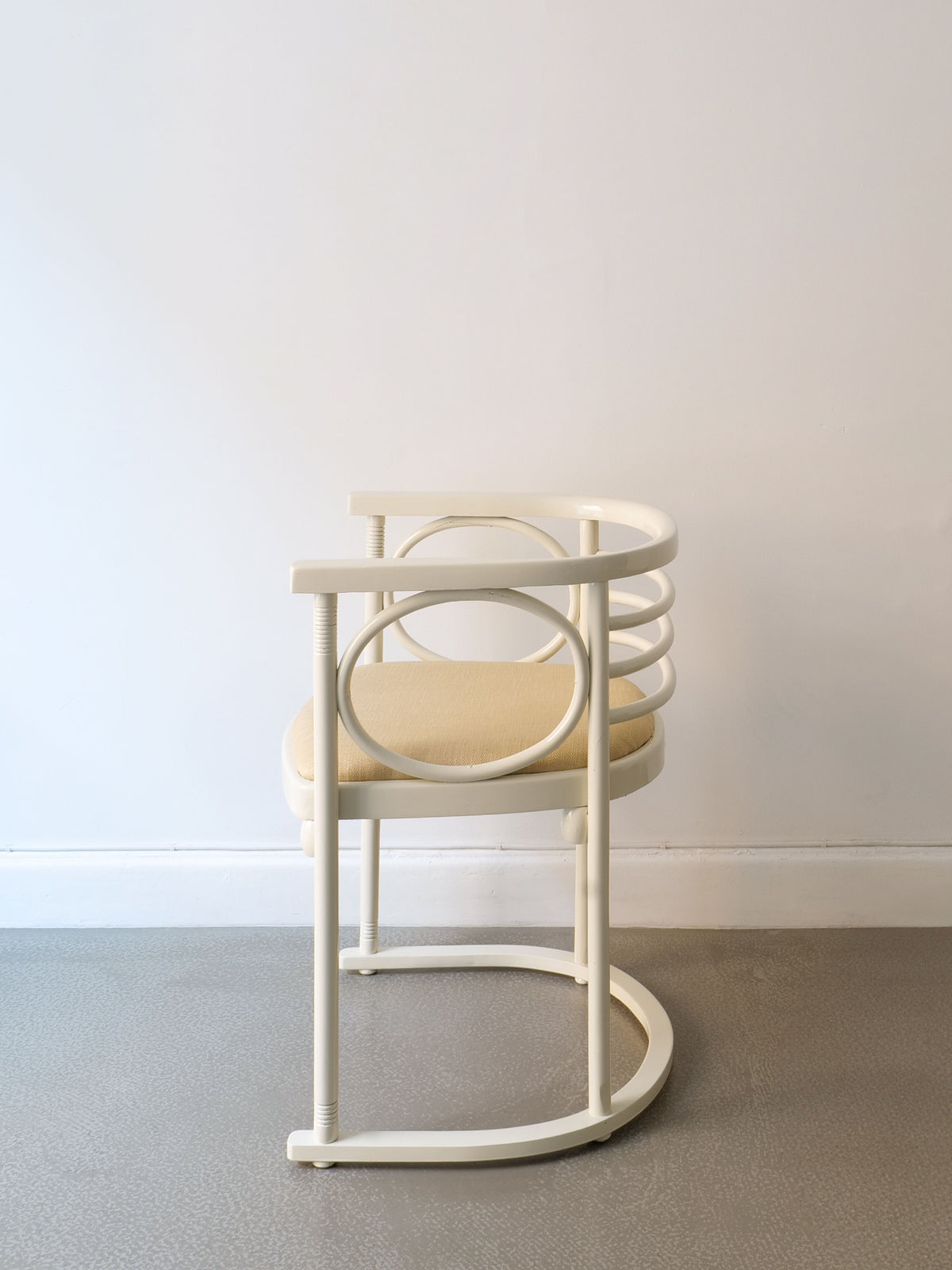 A Thonet Bentwood Chair by Josef Hoffman from Collection apart, featuring a minimalist design with a round backrest and circular side details, stands elegantly against a plain white wall. Its light-colored cushioned seat contrasts beautifully with the textured gray floor.