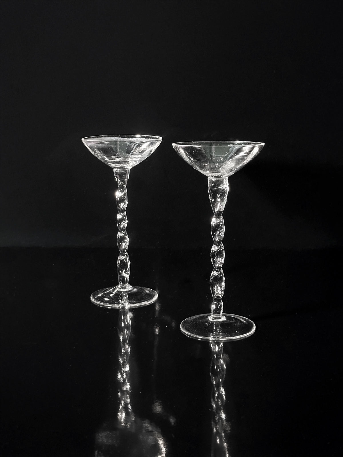 Eight iridescent Art Nouveau liqueur glasses by Dodo Vintage (1910s) are displayed side by side on a reflective black surface, set against a black background.