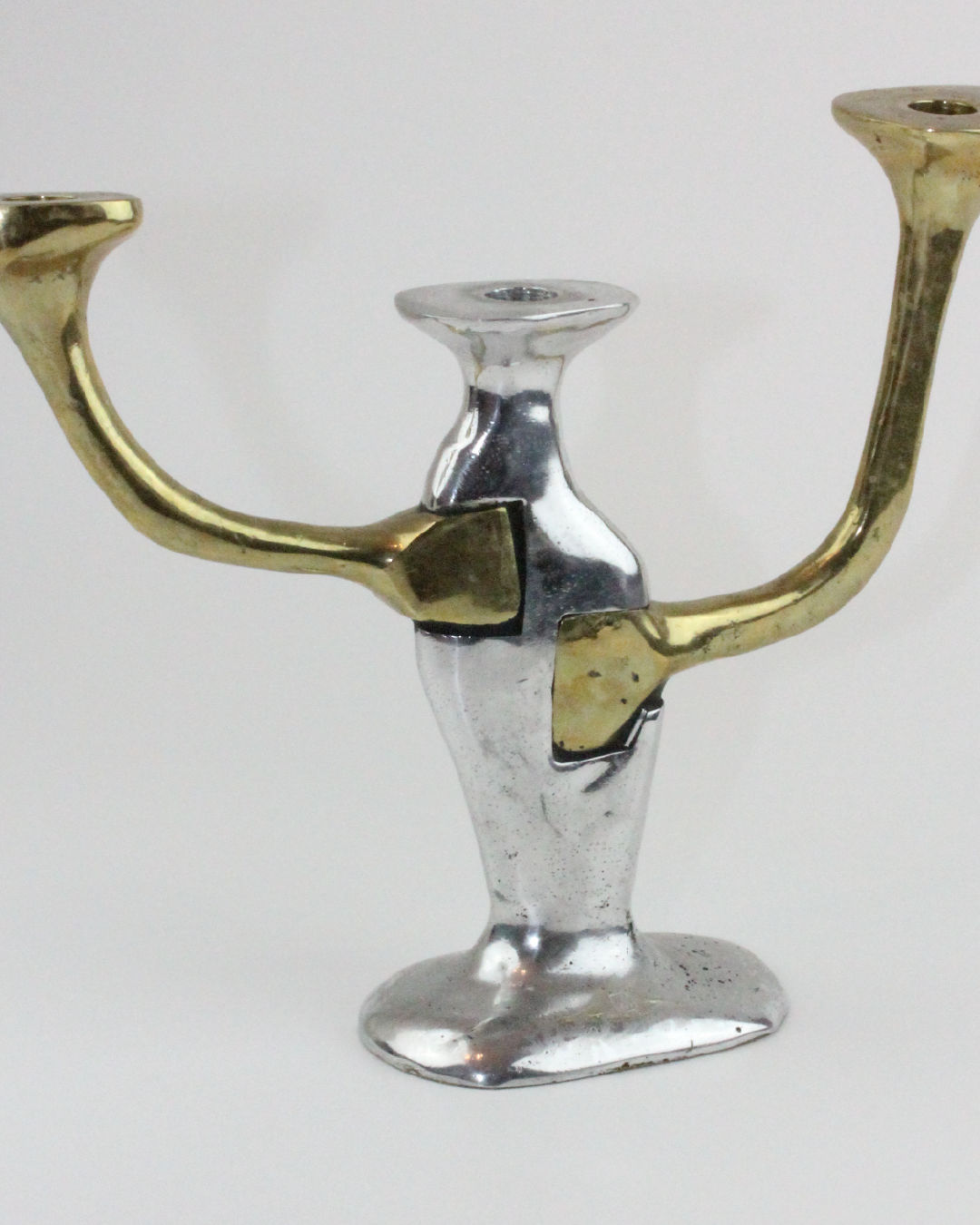 The Brutalist Candle Holder from Boga Avante Shop is a unique piece featuring three arms, with the central stem and base in metallic silver and the two side arms in gold. Its design incorporates abstraction and fluid lines, evoking a 1970s Seville aesthetic that gives it an artistic and modern appearance.