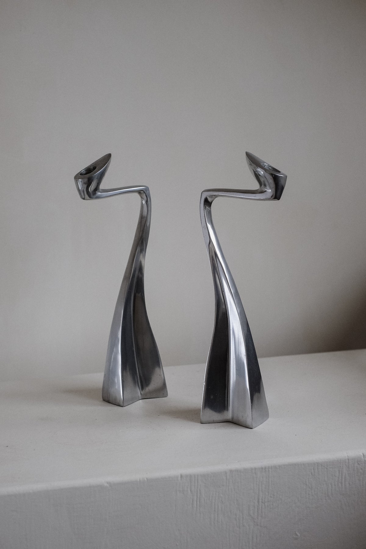 A pair of Matthew Hilton 80's candle holders by septembre studios, with abstract, curvy stainless steel designs, stands on a white surface, exuding a dynamic and modern appearance reminiscent of collector's pieces.