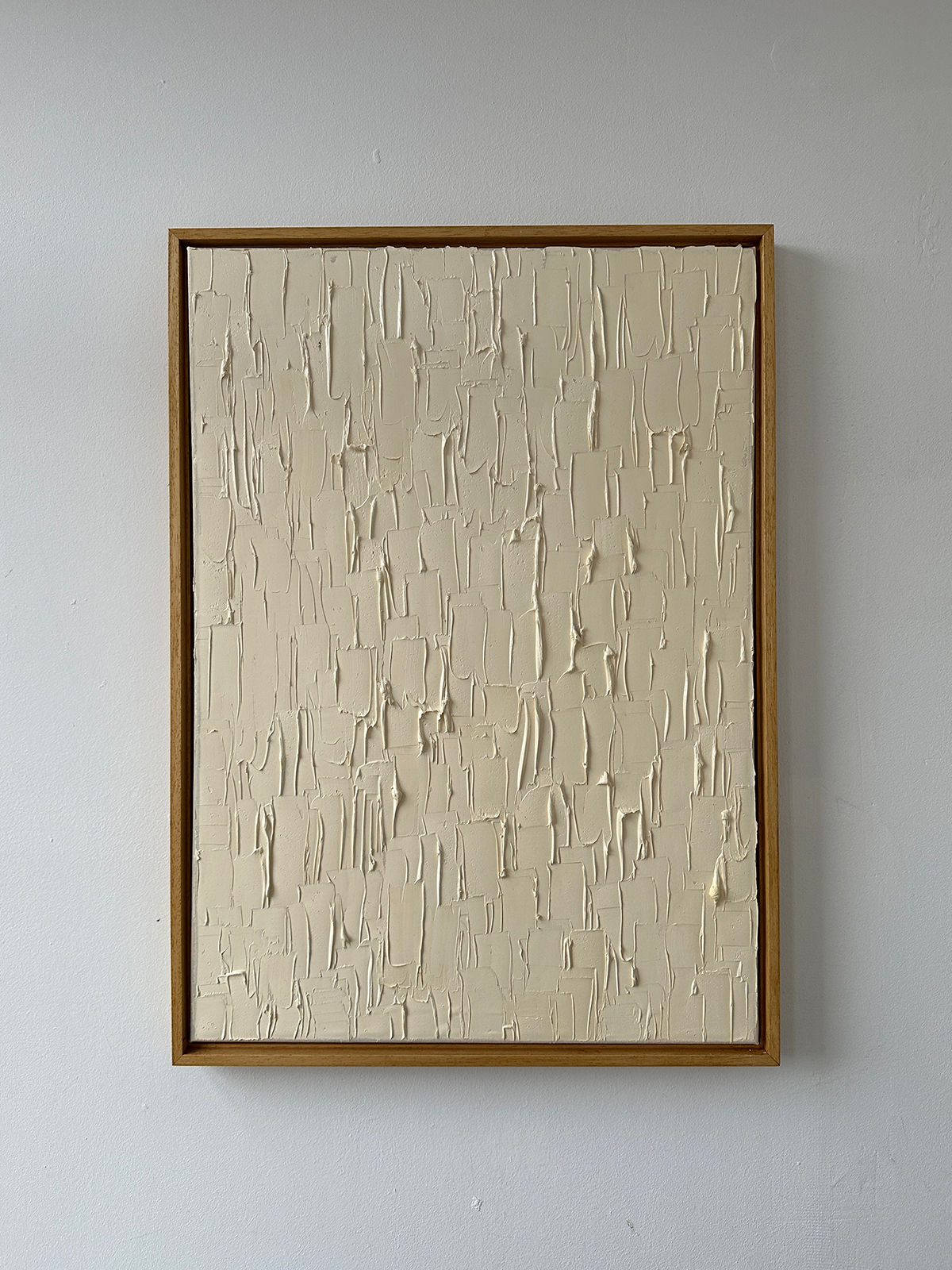 The "Warm White" painting by Roi Elmaliah is a striking framed abstract piece crafted with oil on canvas. It features a textured cream surface adorned with raised, uneven vertical and horizontal patterns, set beautifully against a plain white wall. This captivating artwork echoes the rich art culture of Amsterdam.