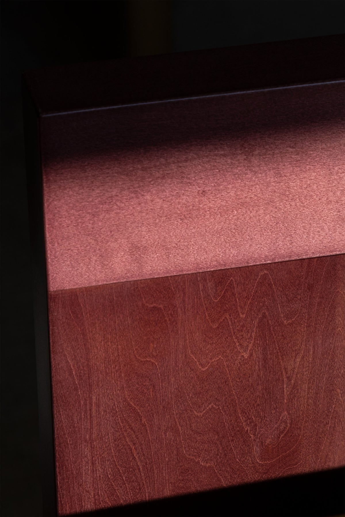 A close-up image of a wooden surface with a rich, dark brown color. The top section is bathed in light, showcasing the wood's texture and grain pattern, while the lower section remains in shadow, emphasizing the contrasting lighting—perfect for highlighting contemporary furniture like the Raja Chair by Juntos Projects.