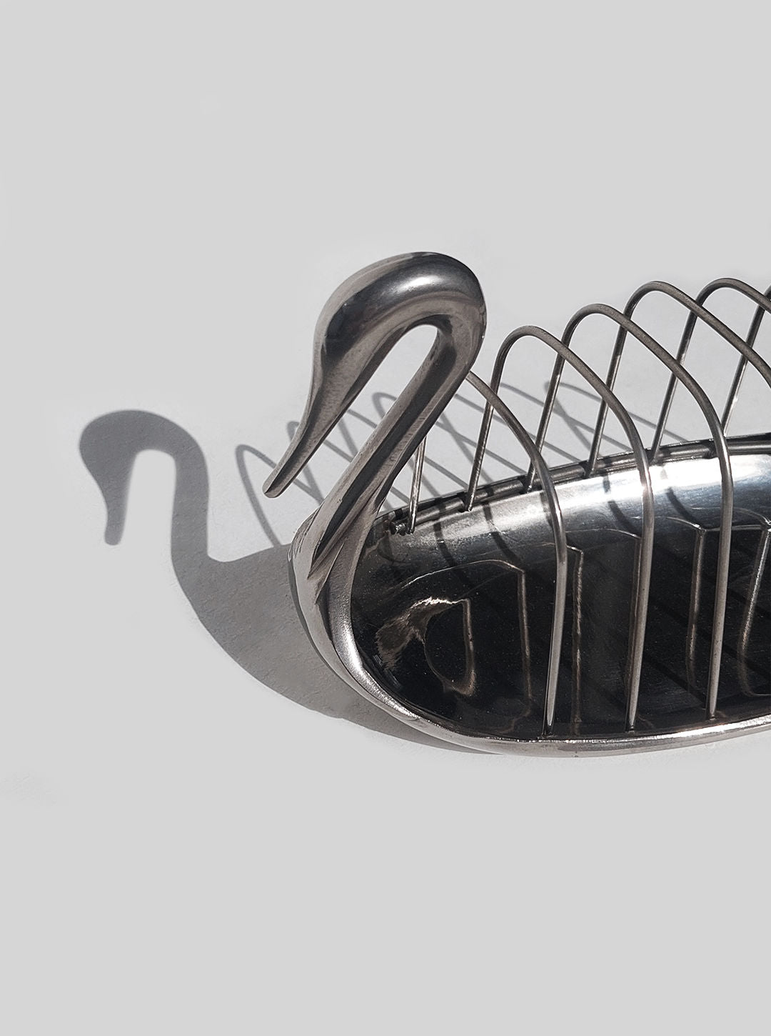 A Les Objoies Swan Toast Rack with a fine mesh basket and an elongated neck forming a graceful handle, casting a faint shadow on a light grey background.