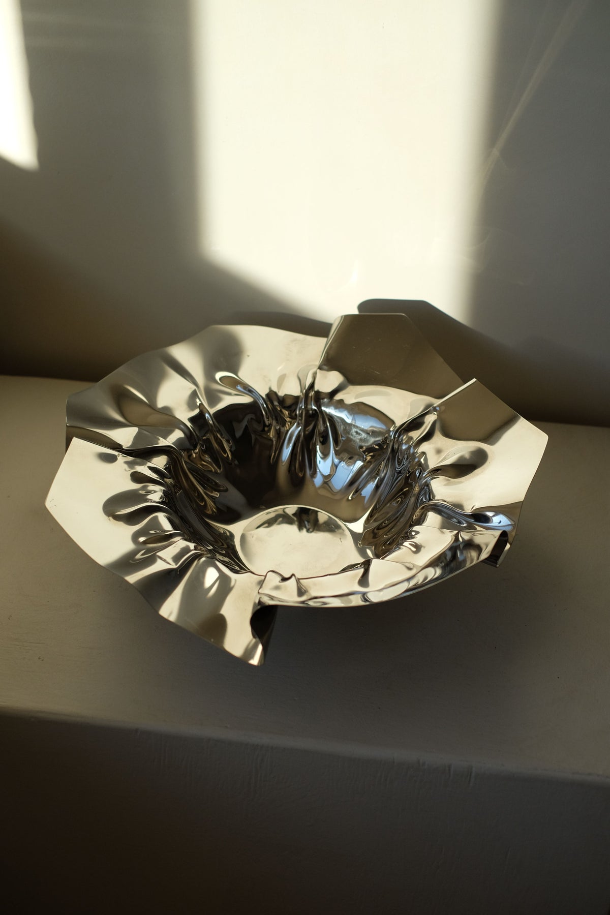 The Folded Chrome Bowl by septembre studios is a shiny, abstract silver piece with a folded shape and crumpled appearance that beautifully reflects light. Resembling a modern art sculpture, it sits elegantly on a white surface, casting soft shadows on the pale wall behind it.