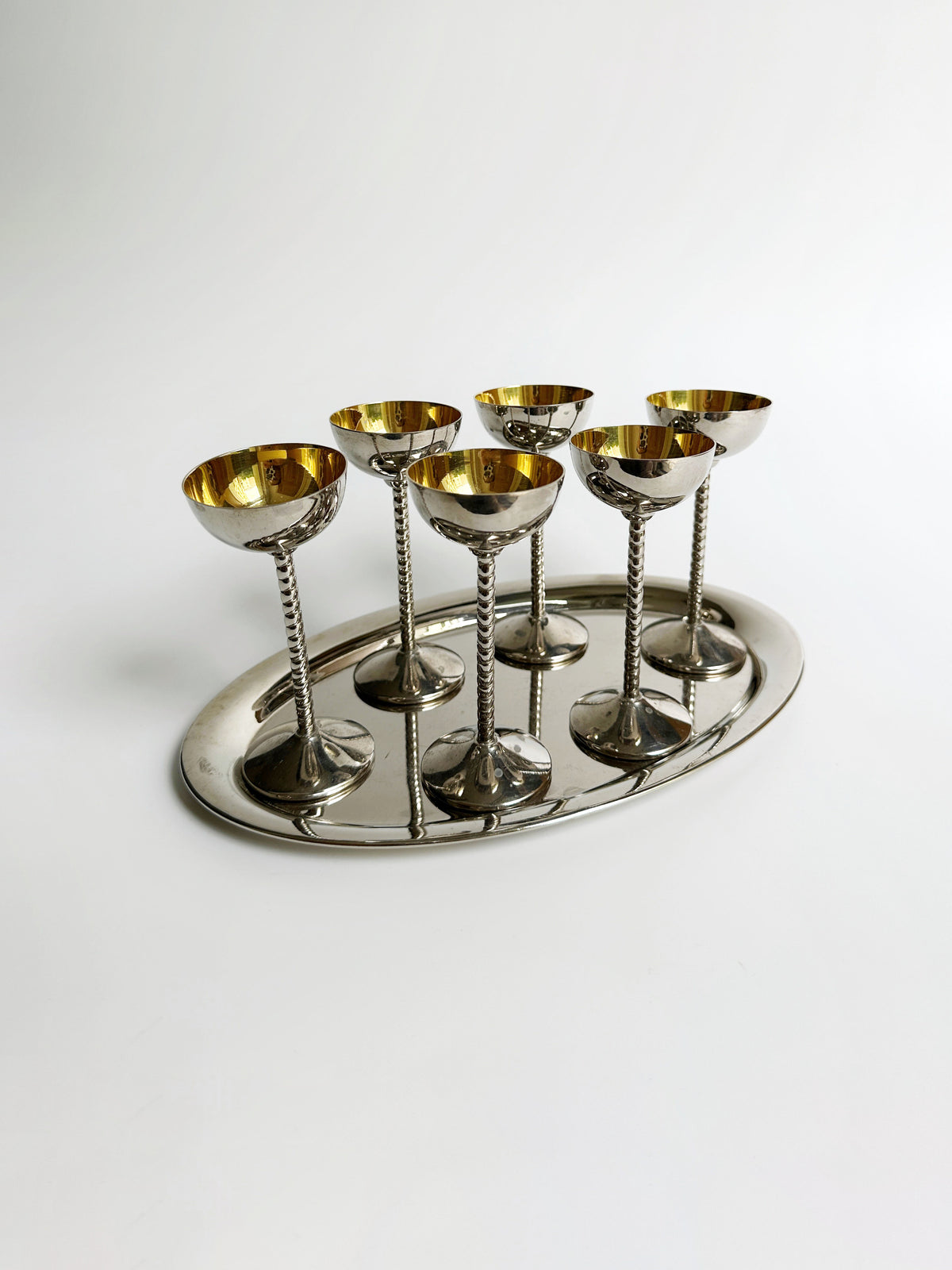 An elegant set of six Dodo Vintage Art Nouveau liqueur goblets with golden interiors, arranged neatly on an elongated silver tray. The goblets feature intricately designed stems that reflect light, showcasing a sophisticated and classic design against a plain white background.