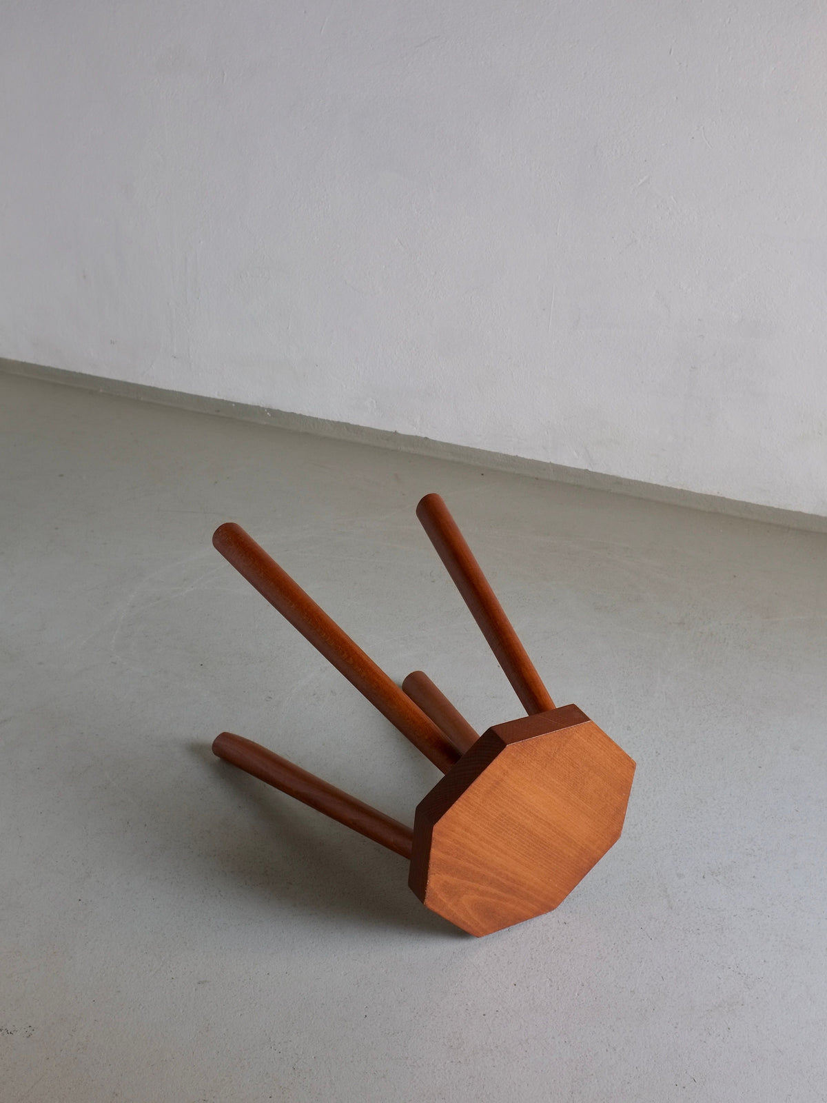 A small, vintage solid wood octagonal stool from the 1960s collection by Veter Vintage is lying on its side on a smooth, light-colored floor in front of a plain, light-gray wall.