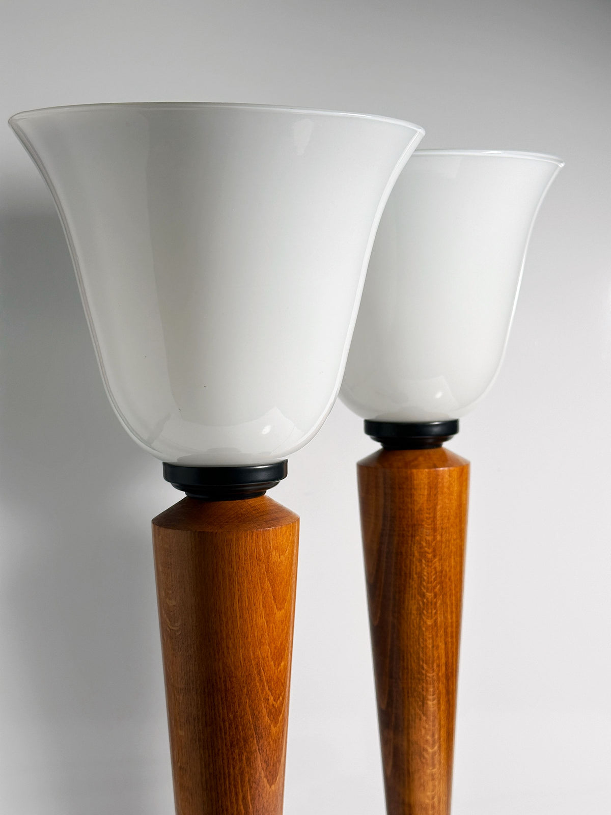 Against a light gray background, two tall floor lamps from Dodo Vintage's "Pair of Art Deco Chalices with Teak Wood" boast frosted glass shades and dark wood bases, blending smooth glass with rich wood in an elegant, modern contrast.