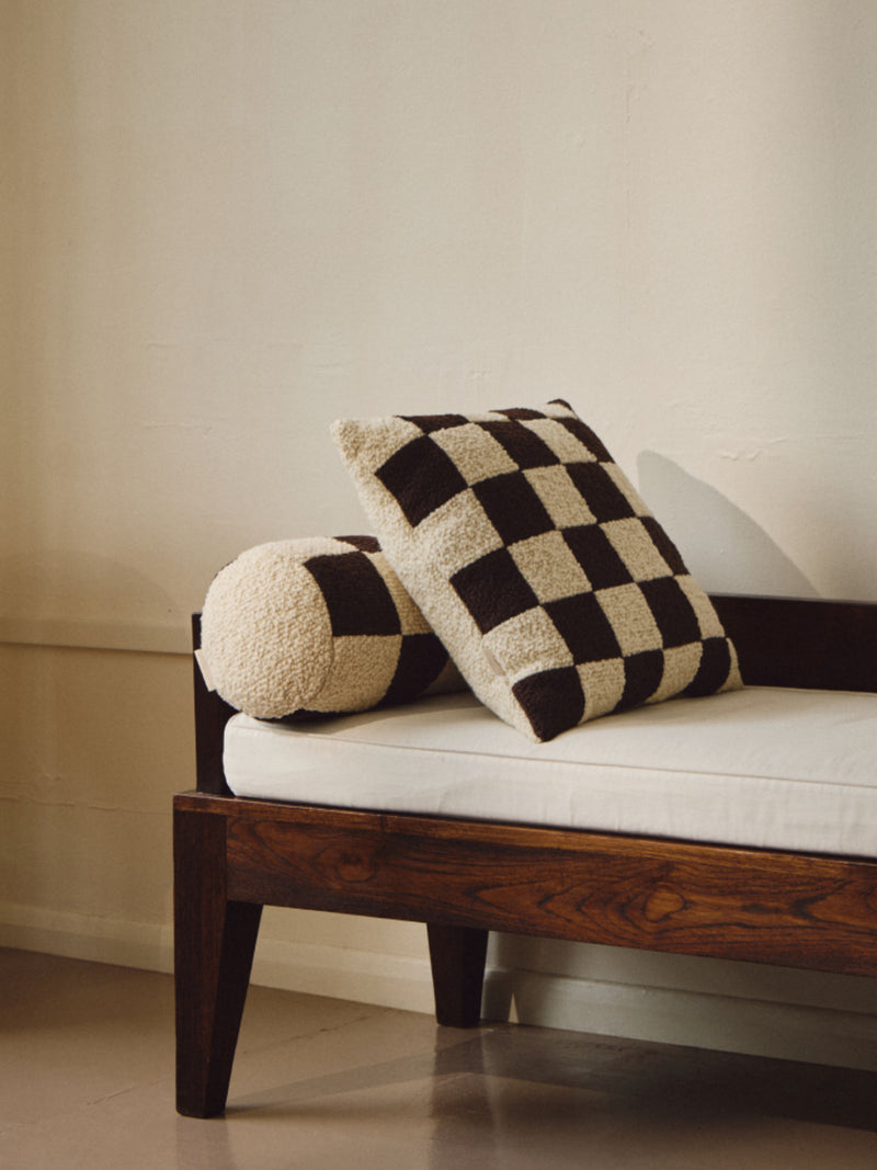 A dark wooden bench with a white cushion is adorned with two pillows: one rectangular with a black and white checkered pattern and another cylindrical one textured in cream. The Le Carré - Wool Bouclé - Chocolate/Eggshell by AMÉLIORÉ features British eco wool, while the neutral-colored wall in the background enhances the minimalist aesthetic.