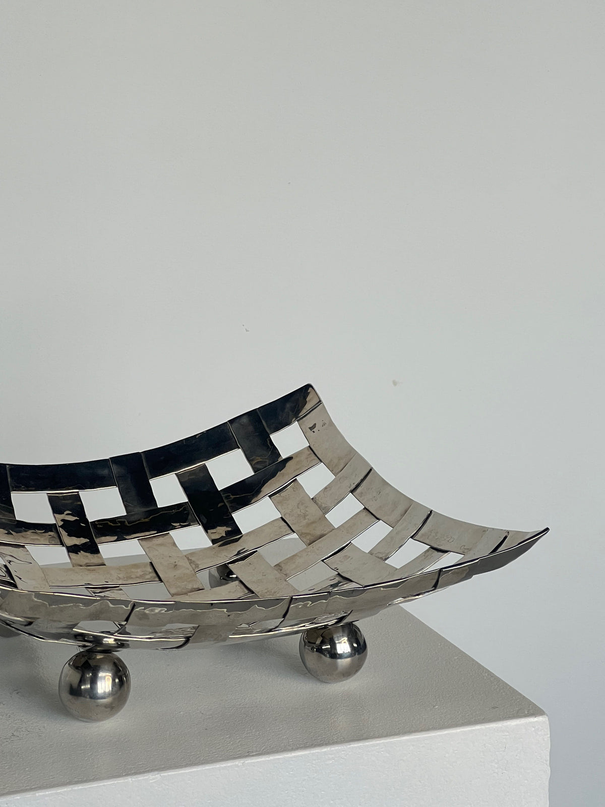 The Aliminum Dish on Feet by Porter Studio is a metallic woven basket with a sleek polished aluminum finish. It elegantly sits on a small pedestal, featuring two shiny spherical ball feet, set against a plain gray background.