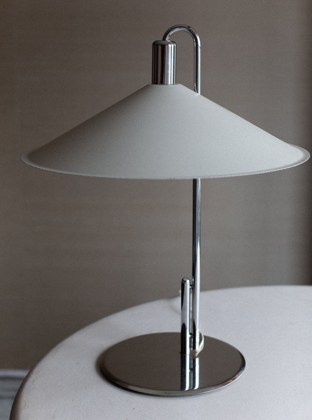 A modern Börge Lindau Lamp by Out For Lunch stands on a round white surface, embodying a minimalist aesthetic. It features a sleek, chrome base and a slender upright stem. The conical, light-colored fabric lampshade has a slight tilt, casting soft light upwards and downwards. The background is neutral, enhancing its Scandinavian design.