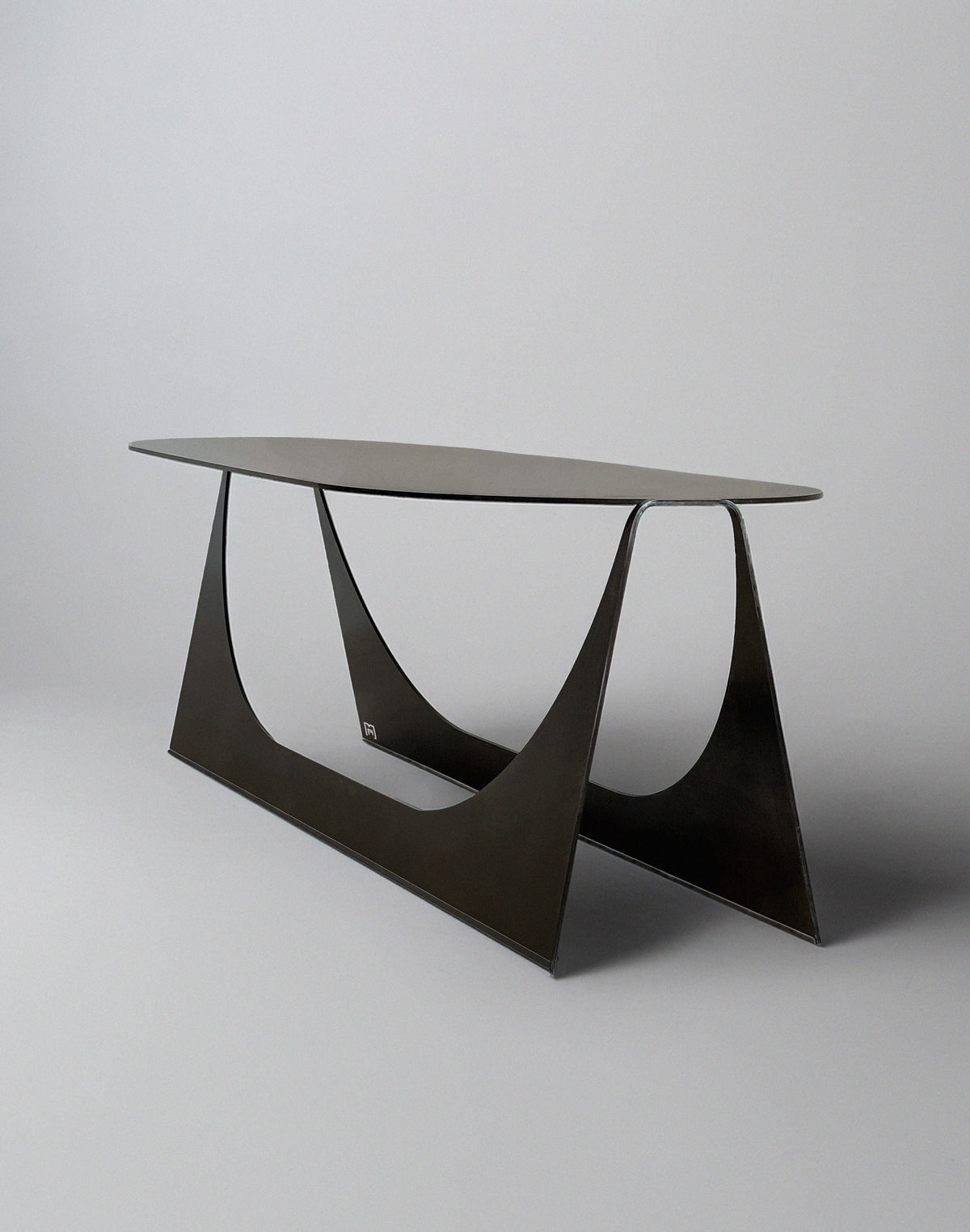 The 'Tani' Coffee Table by Modan is a modern, minimalist oval table in black steel with a slim top and geometric triangular base, shown against a plain light gray background.