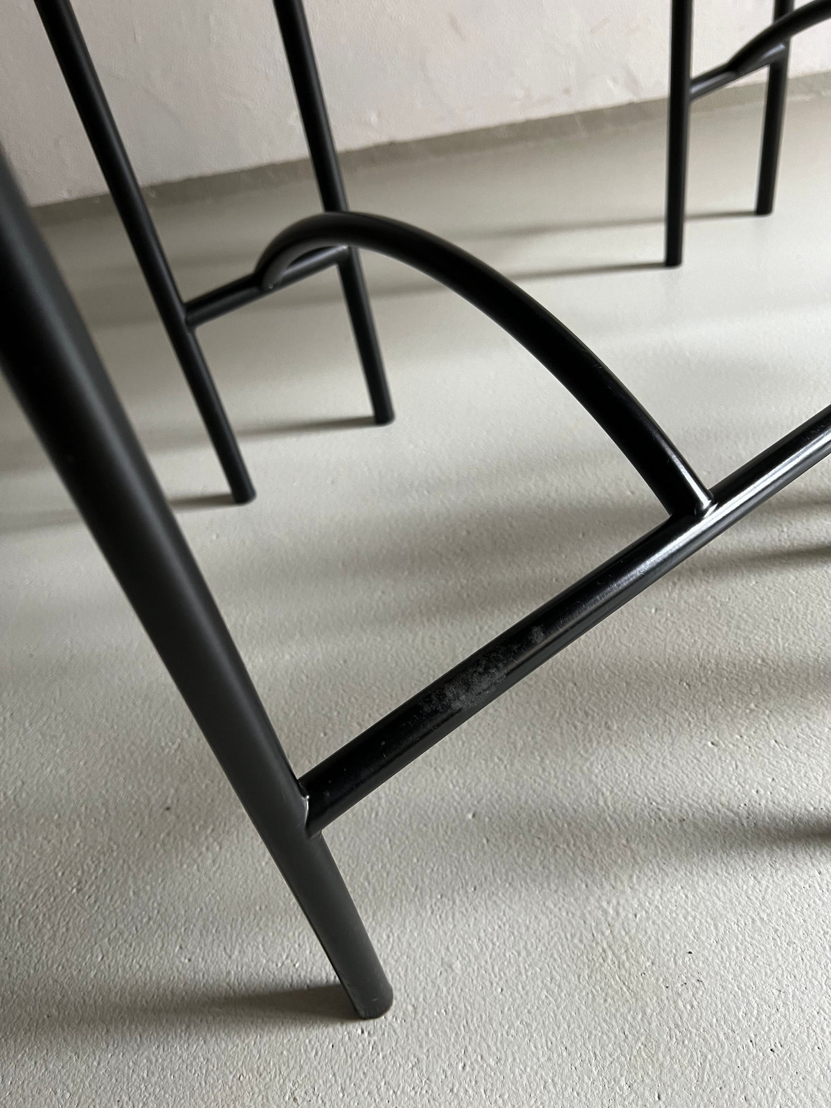 Close-up of a black metal furniture leg with a curved support piece, positioned on a light-colored floor. The design features sleek, straight lines and minimalist aesthetics, creating a modern and clean look that complements the faux leather upholstery of 4 Postmodern "Tokyo" Chairs by Rodney Kinsman for Veter Vintage | Italy | 1980s.