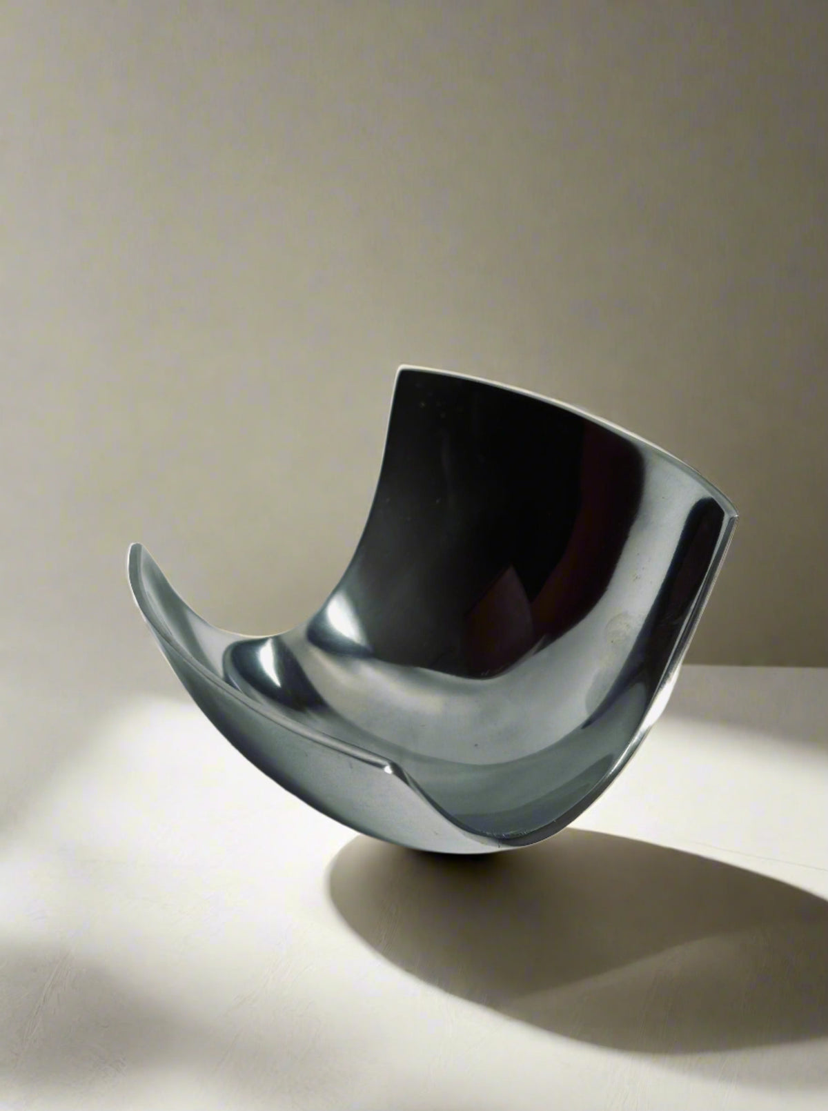 A Modernist Decorative Dish with a unique curved shape crafted from steel by Flock Studio rests on a light-colored surface, casting a soft shadow. Its smooth, reflective finish contrasts beautifully with the neutral background.
