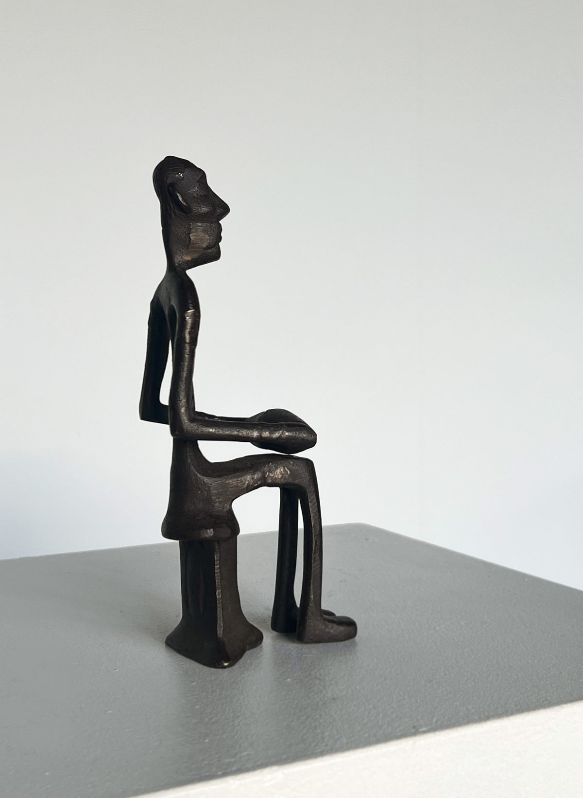 The Bronze Drummer Man Sculpture by Porter Studio features a minimalist abstract figure with elongated limbs and angular features, seated on a stool. Its precise bronze design creates a dynamic rhythm against a plain, light background.