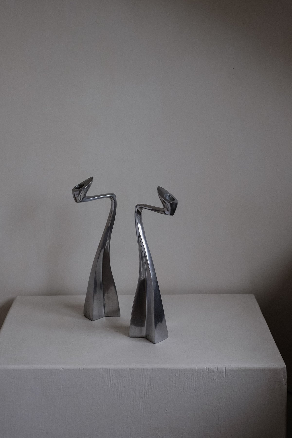 A pair of sleek, modern metal candleholders by Matthew Hilton from the 80s, offered by septembre studios, rest elegantly on a white pedestal against an off-white wall. These collector's pieces boast abstract curving forms with smooth, reflective surfaces that evoke grace and sophistication.